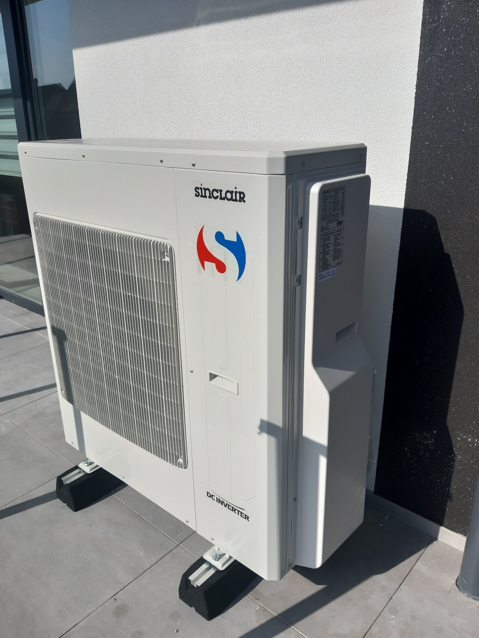 Biała, zewnętrzna jednostka klimatyzacji Sinclair DC Inverter, zamontowana na czarnych podkładkach antywibracyjnych, stoi na szarym, betonowym podłożu przy białej ścianie budynku.