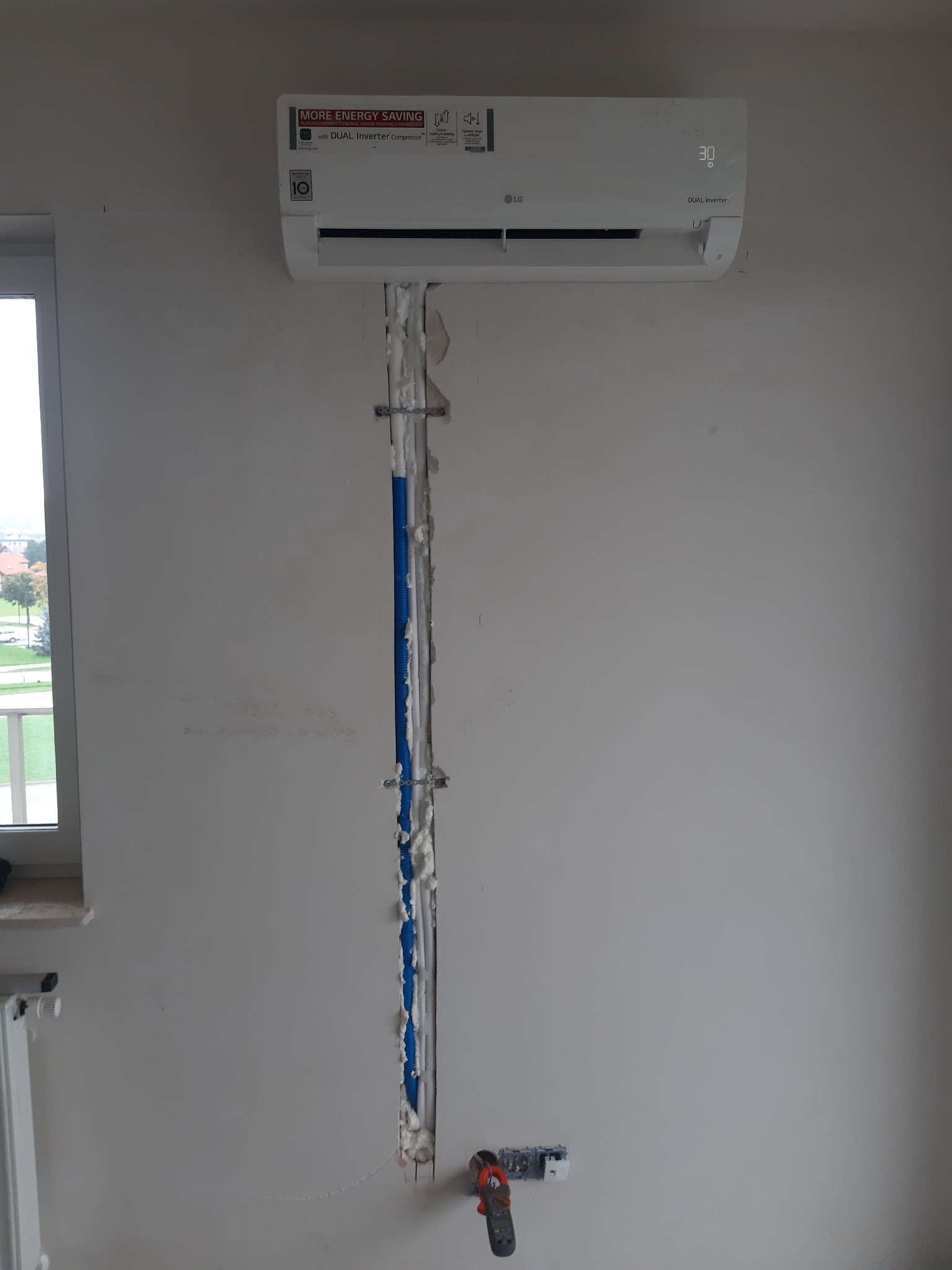 Montaż klimatyzatora LG Dual Inverter na ścianie, widoczne nieukończone prace instalacyjne z niebieskimi przewodami i pianką montażową w bruździe ściennej, obok okna i grzejnika.