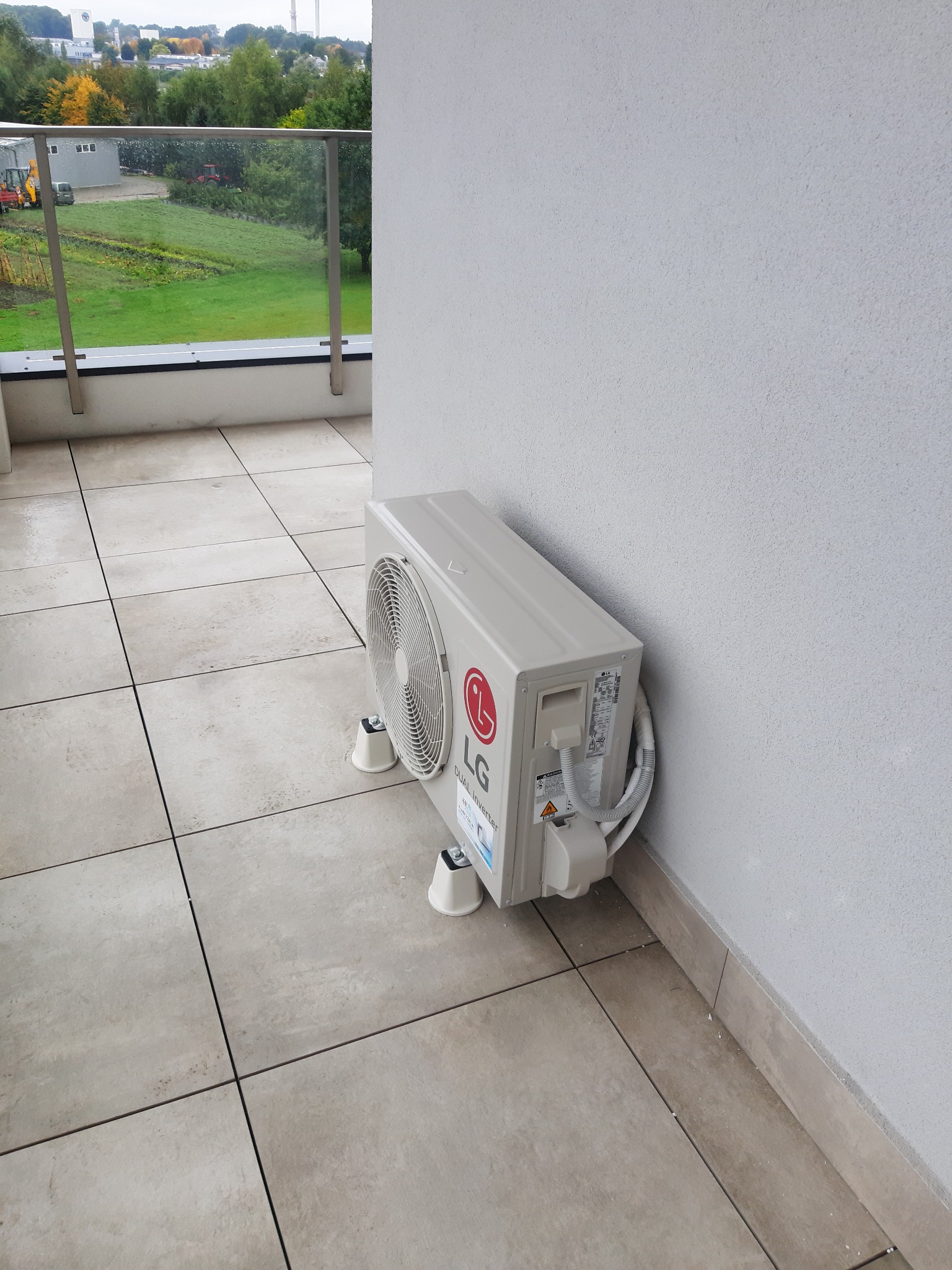 Zewnętrzna jednostka klimatyzacji LG Dual Inverter umieszczona na balkonie z widokiem na tereny zielone i budynki w tle.