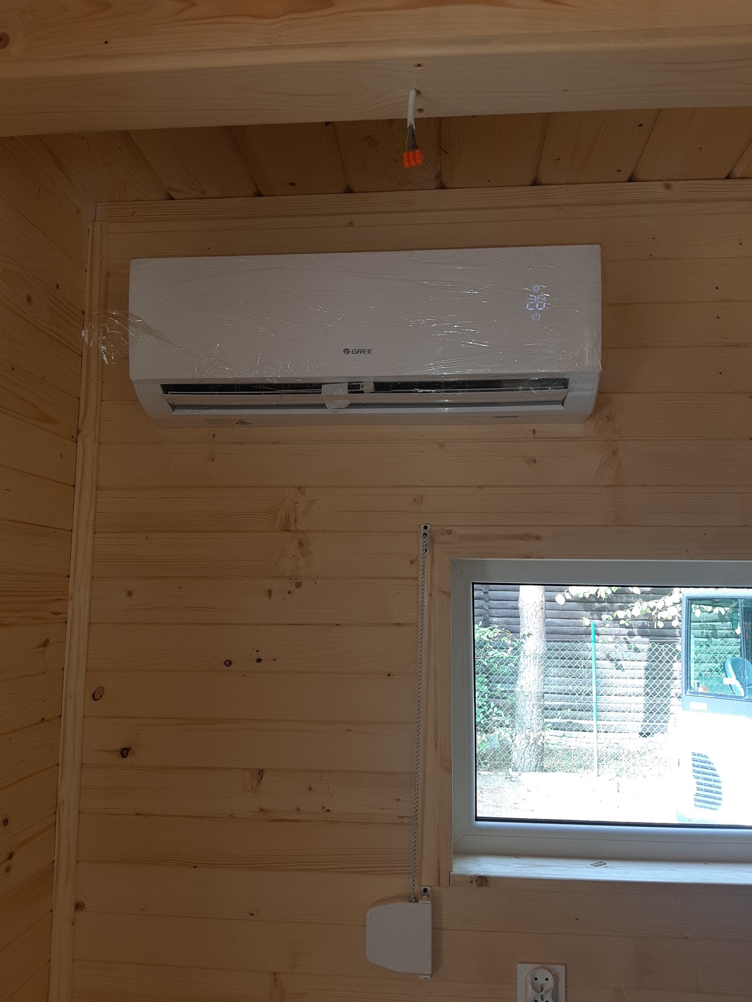 Nowo zamontowany klimatyzator marki Gree, jeszcze w folii ochronnej, na drewnianej ścianie wewnątrz pomieszczenia, widoczne okno z widokiem na zewnątrz.