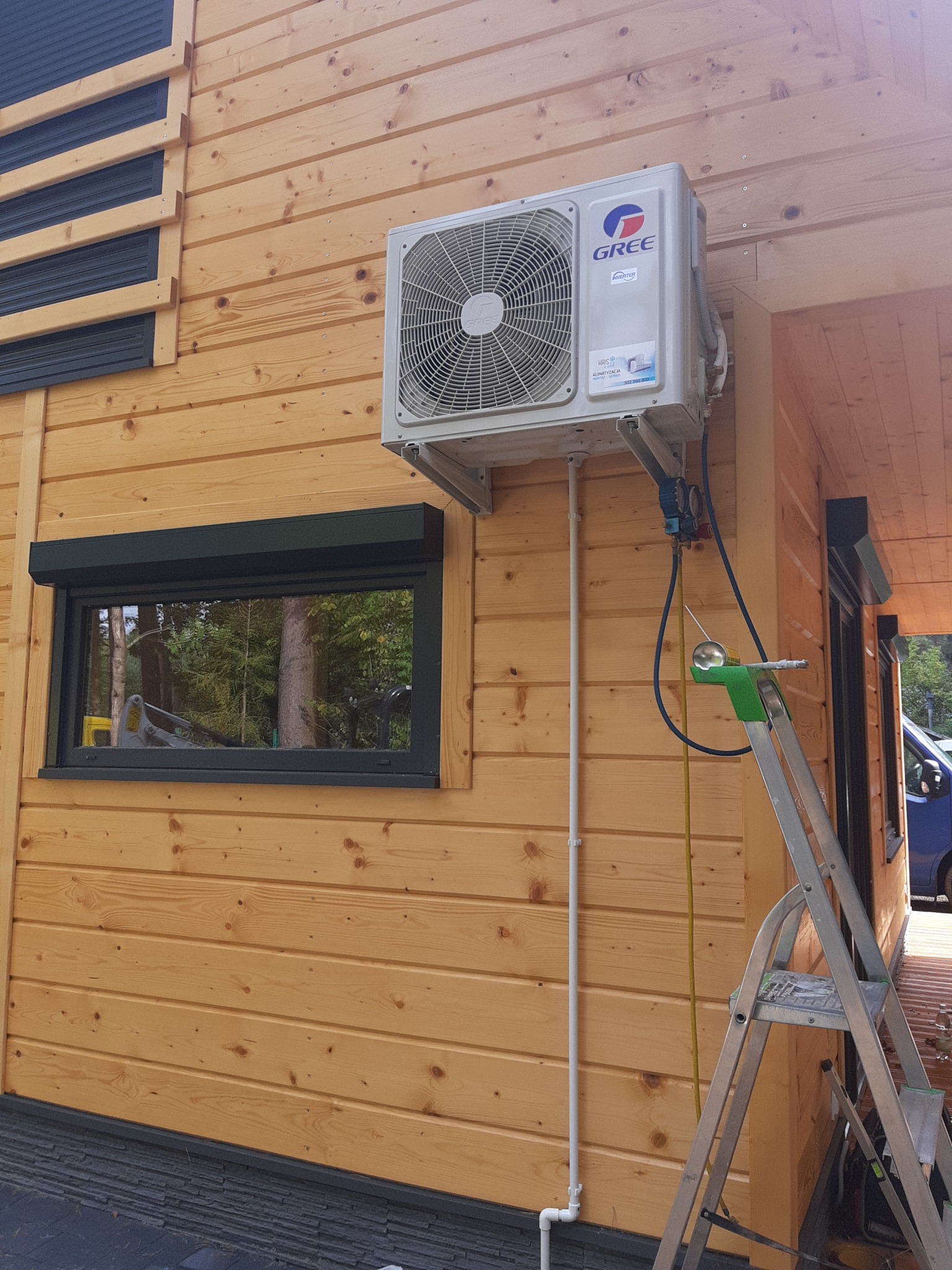 Zamontowana jednostka zewnętrzna klimatyzacji Gree na drewnianej elewacji domu, widoczne rury i przewody, drabina oparta o ścianę, narzędzia serwisowe.