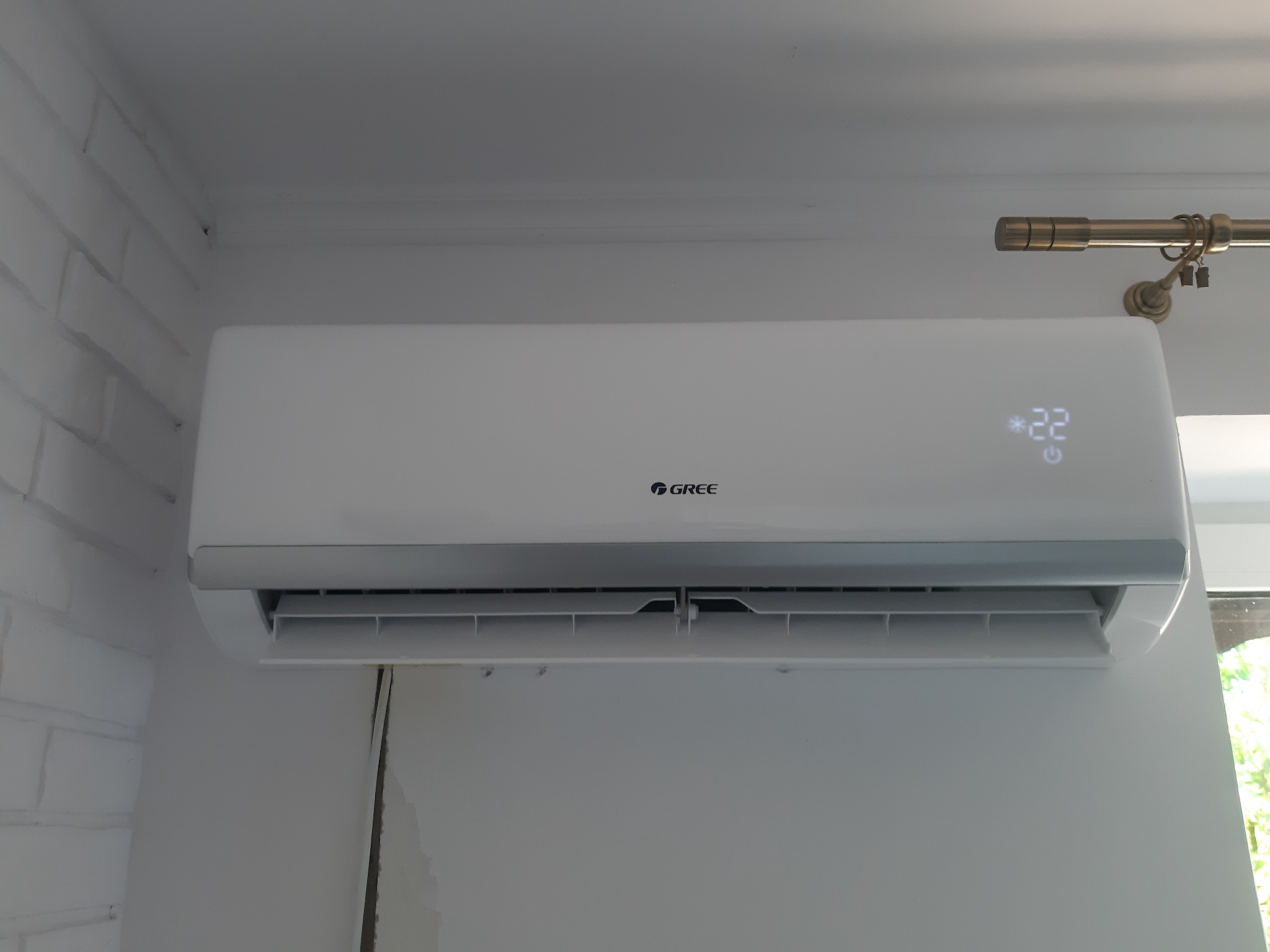 Biała klimatyzacja marki Gree zamontowana na ścianie, ustawiona na 22 stopnie, widoczna część okna i karnisza.