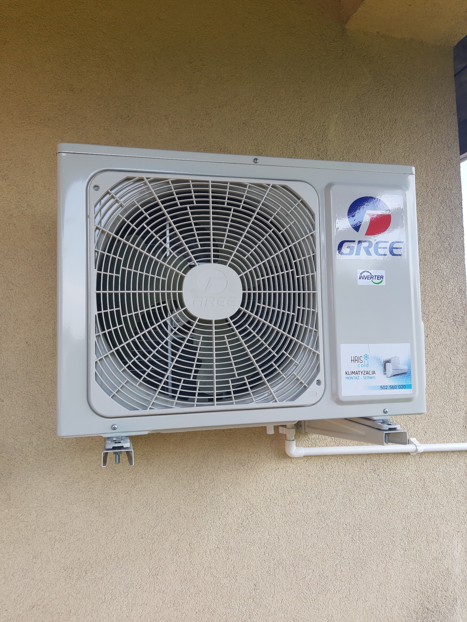 Zewnętrzna jednostka klimatyzacji marki Gree zamontowana na elewacji budynku, widoczne metalowe wsporniki i rury odprowadzające skropliny, naklejka z danymi kontaktowymi firmy montującej.