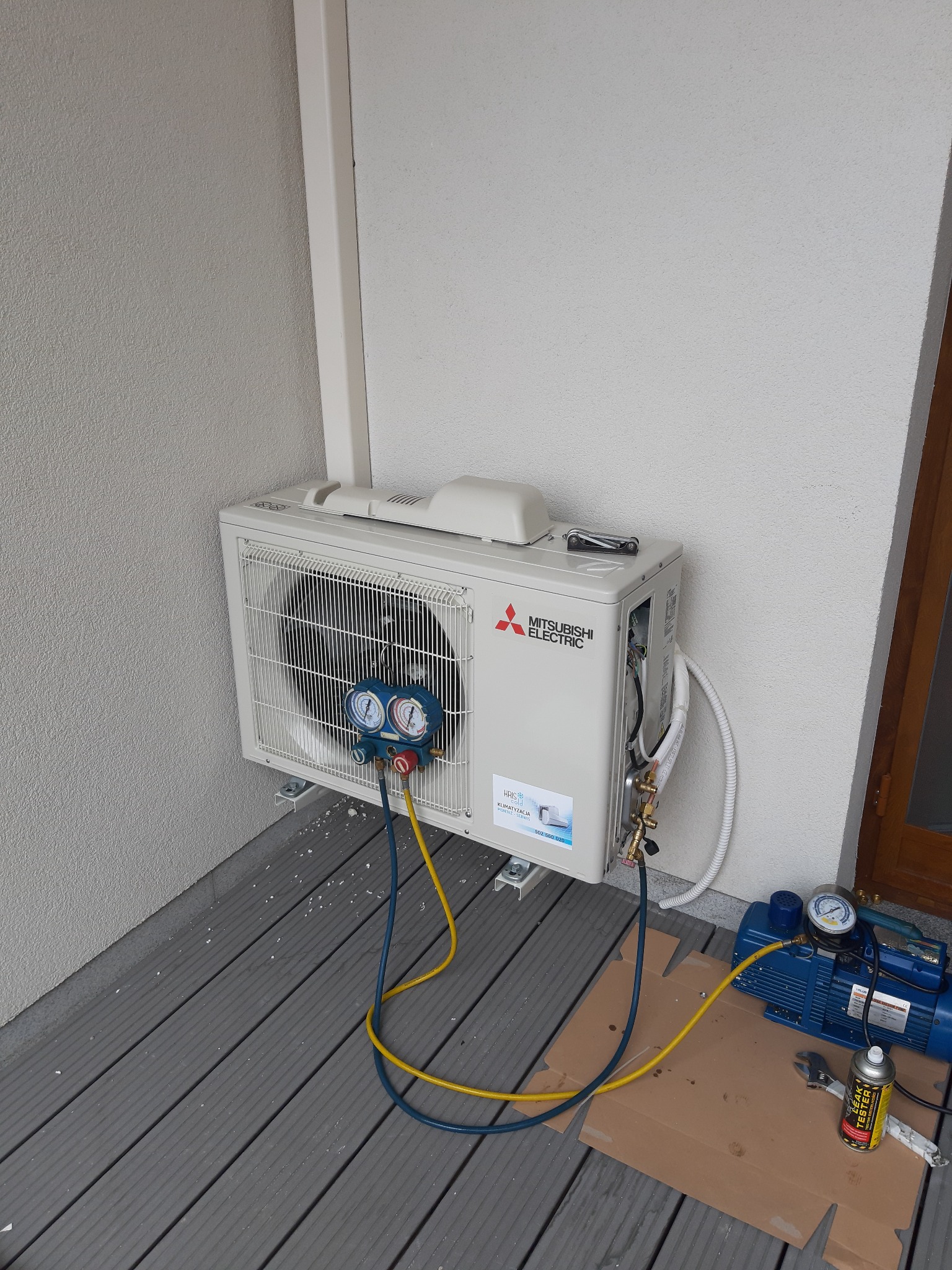 Zewnętrzna jednostka klimatyzacji Mitsubishi Electric zamontowana na ścianie budynku, podłączona do manometrów i pompy próżniowej podczas serwisu, widoczny tester szczelności.