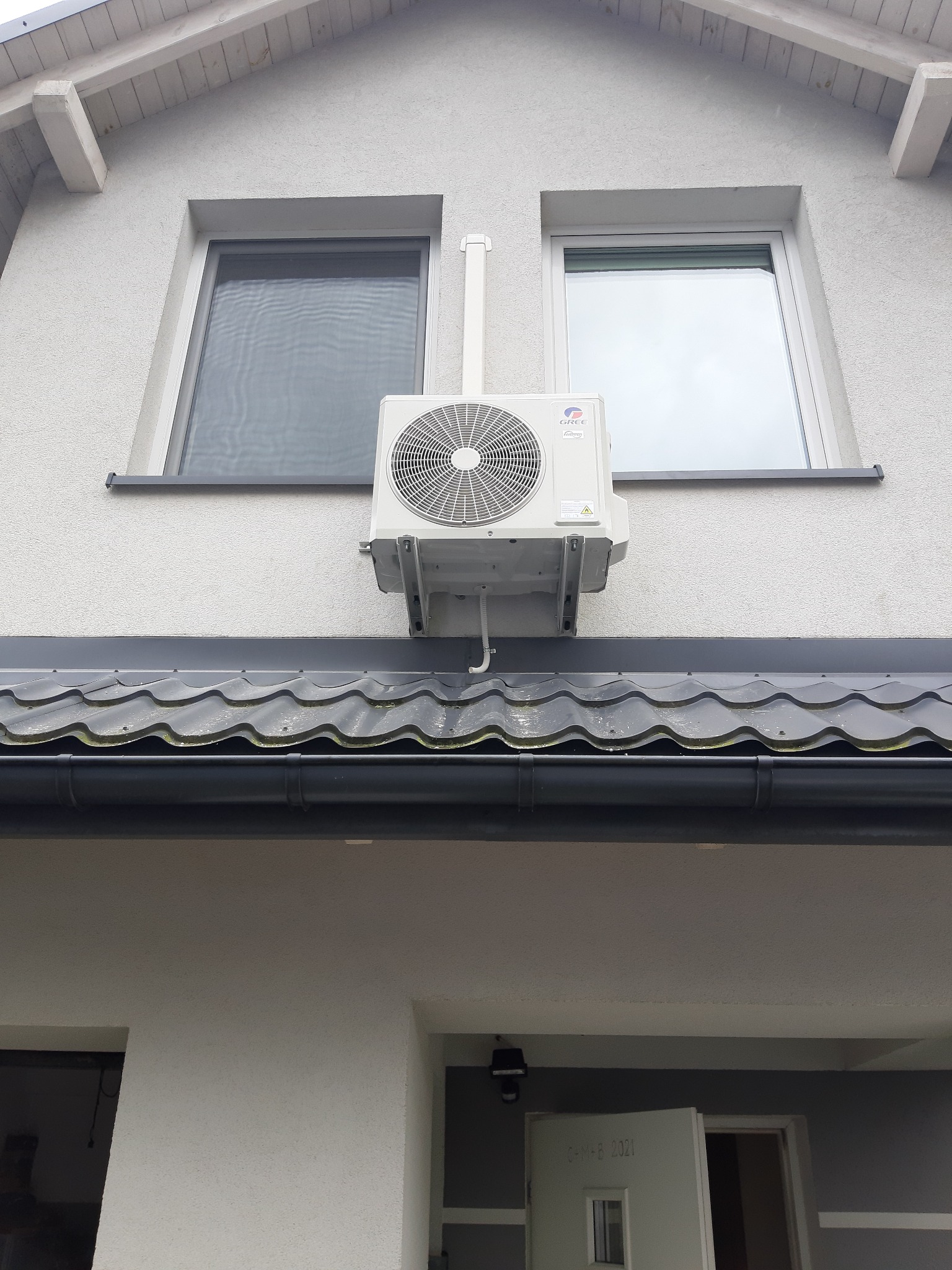 Zewnętrzna jednostka klimatyzacji marki Gree zamontowana na elewacji domu pomiędzy dwoma oknami, widoczna instalacja odprowadzająca skropliny.