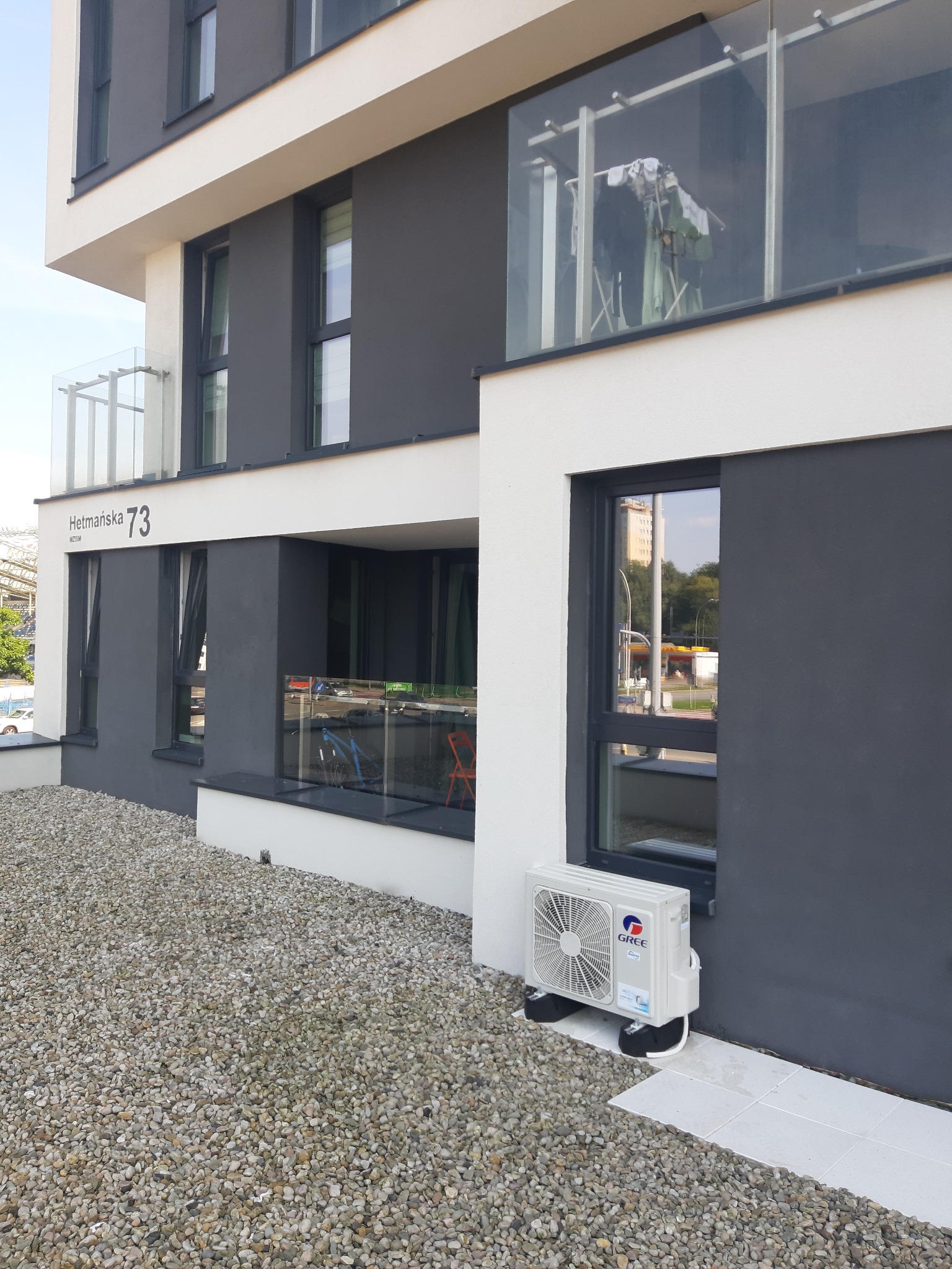 Biała jednostka zewnętrzna klimatyzatora GREE zamontowana na czarnych podkładkach, przy szarej ścianie budynku mieszkalnego z adresem Hetmańska 73, na tle żwirowego podłoża.