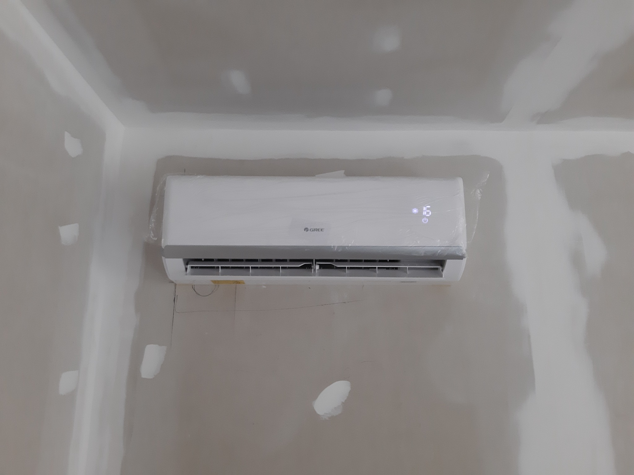 Świeżo zamontowany klimatyzator marki Gree, jeszcze w folii ochronnej, z wyświetlaczem temperatury 16 stopni, na tle ściany w trakcie wykańczania, z widocznymi łatami gipsowymi.