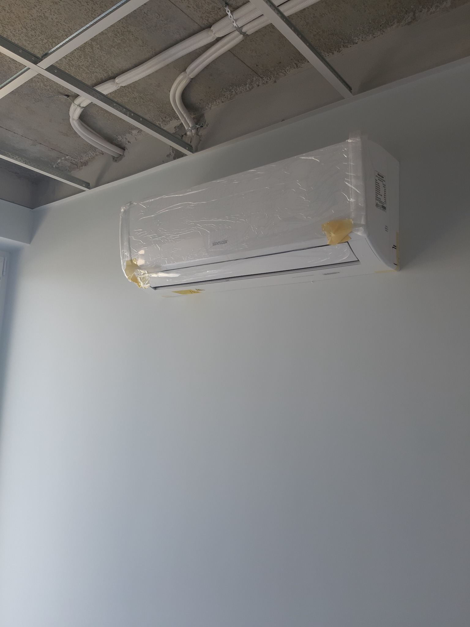 Nowo zamontowany, biały klimatyzator marki Sinclair na jasnoniebieskiej ścianie, jeszcze w folii ochronnej, z widocznymi rurami instalacyjnymi pod sufitem w szarej kratownicy.