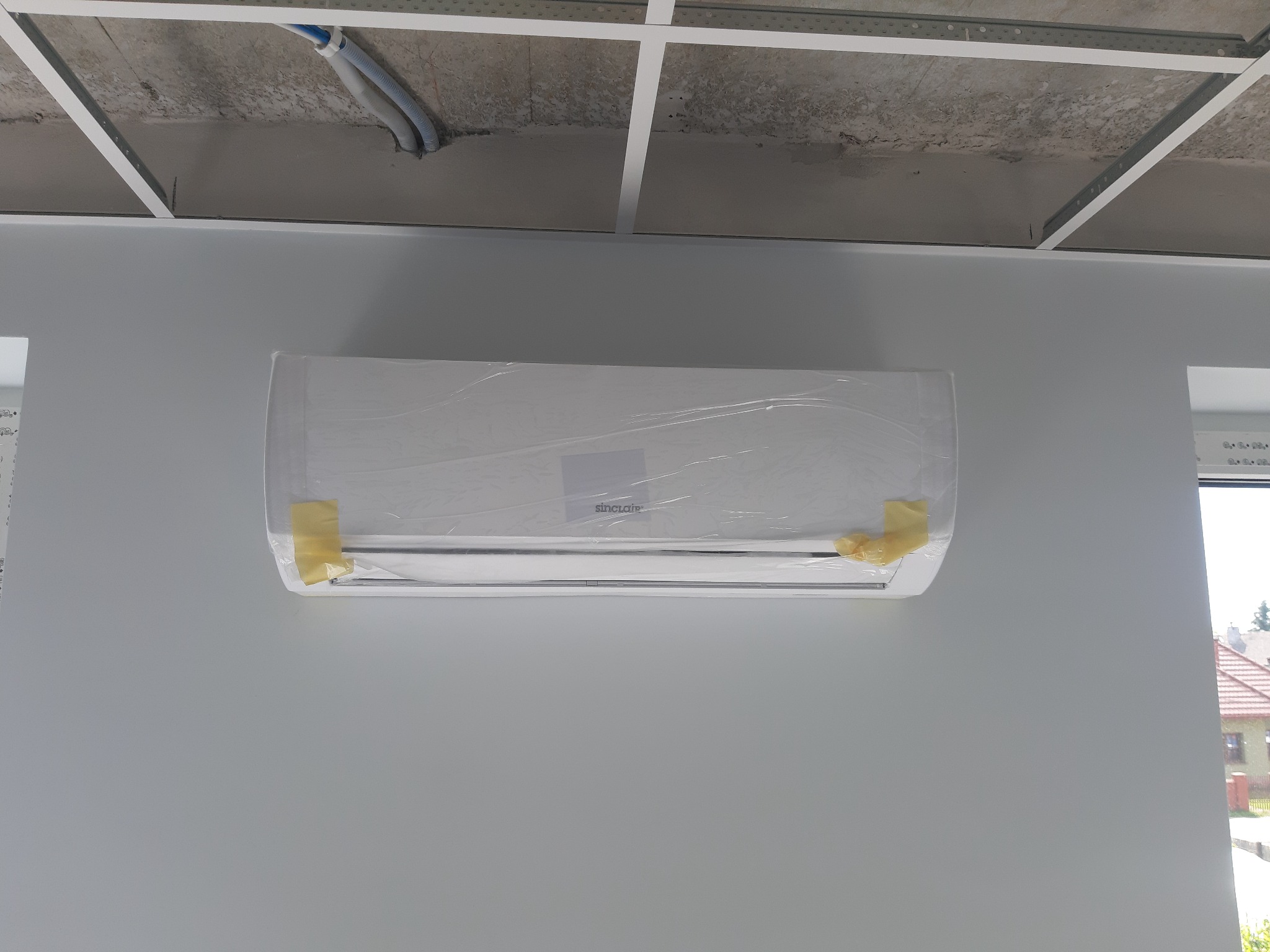 Nowy klimatyzator Sinclair zamontowany na szarej ścianie, zabezpieczony folią i taśmą, widok na sufit podwieszany i okno z widokiem na dom.