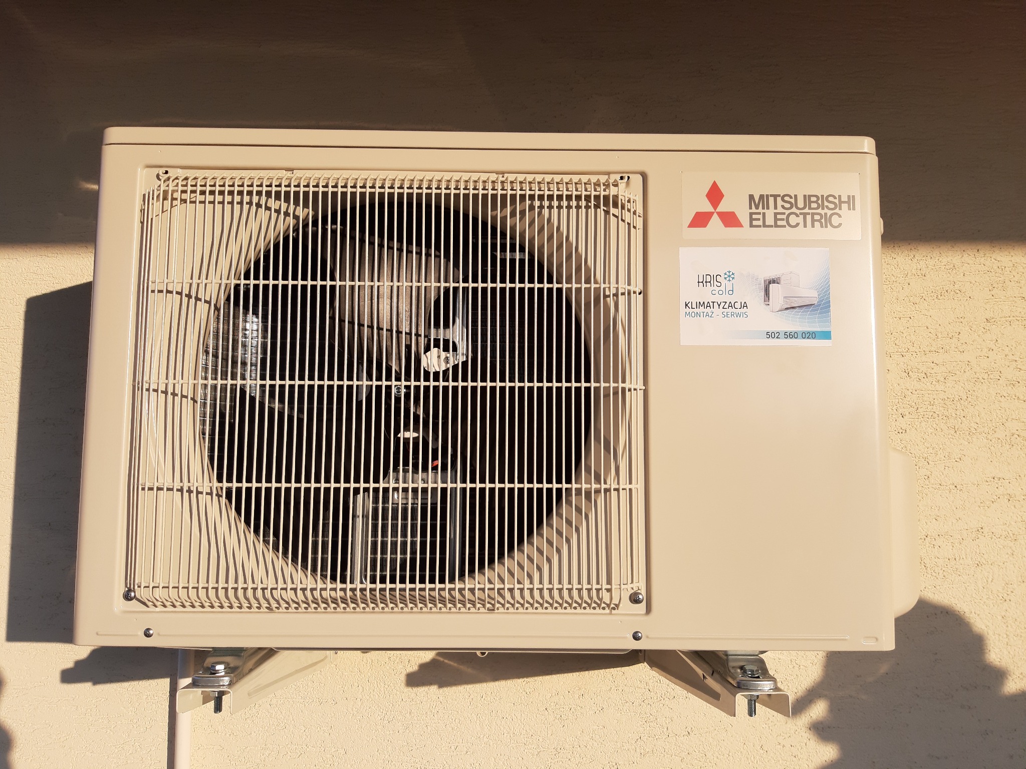 Zewnętrzna jednostka klimatyzacji Mitsubishi Electric zamontowana na ścianie budynku, z naklejką firmy zajmującej się montażem i serwisem.