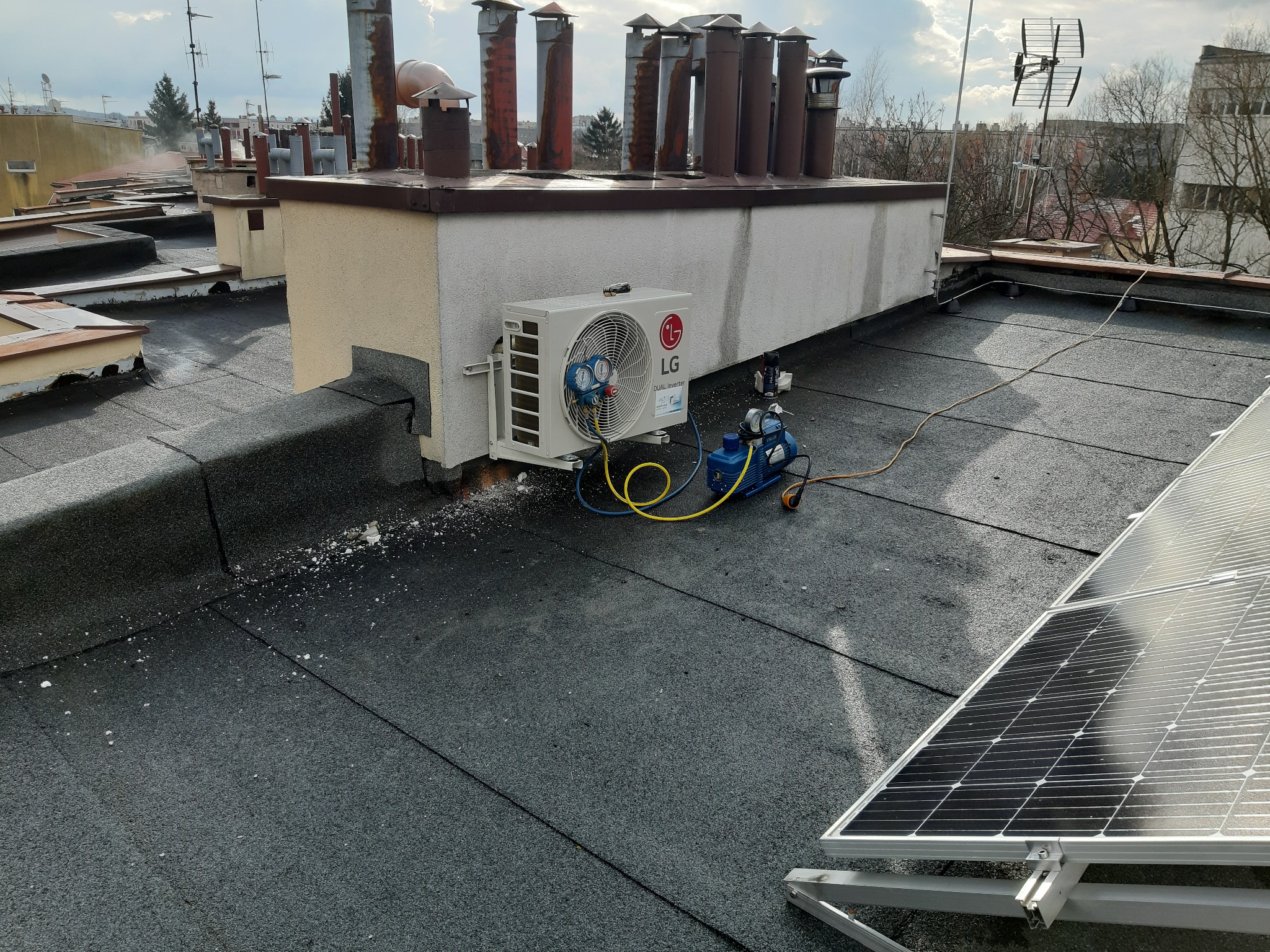 Zewnętrzna jednostka klimatyzatora LG zamontowana na dachu budynku, podłączona do pompy próżniowej, obok panel słoneczny, widoczne przewody i narzędzia.
