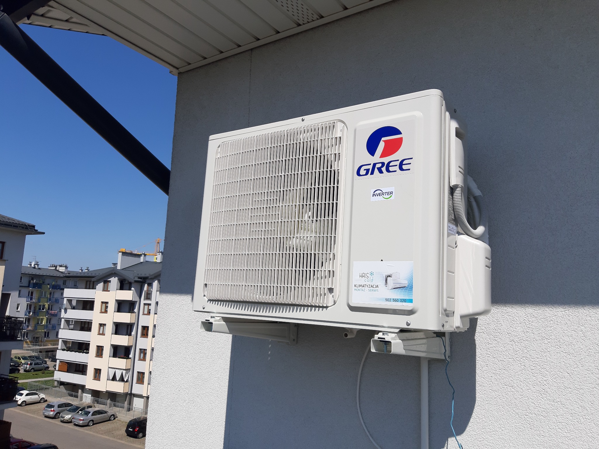 Biała jednostka zewnętrzna klimatyzatora marki Gree zamontowana na elewacji budynku mieszkalnego, z widocznym logotypem firmy instalacyjnej i numerem telefonu.