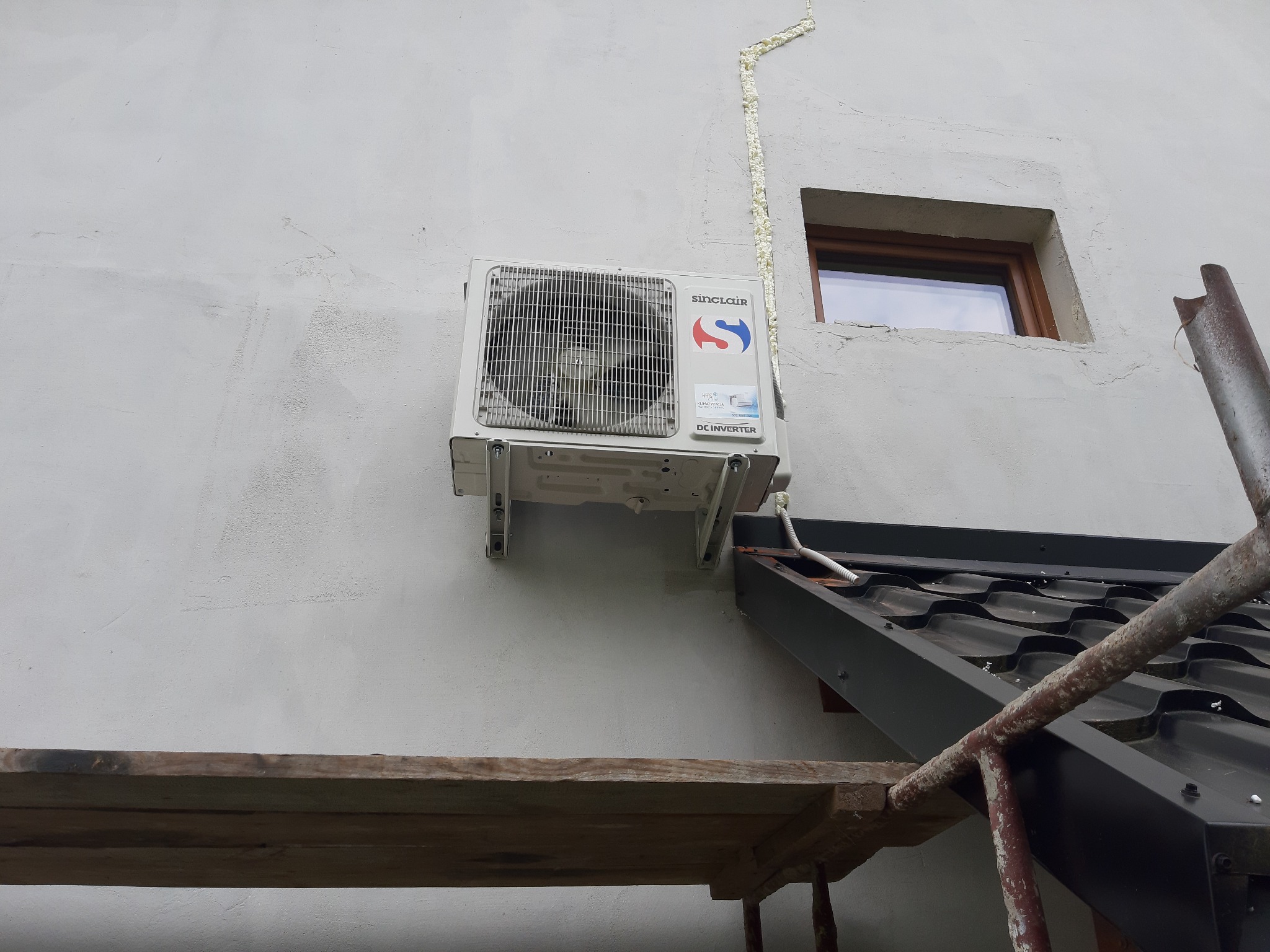 Biała jednostka zewnętrzna klimatyzatora Sinclair zamontowana na ścianie budynku obok okna, z widocznymi śladami pianki montażowej i fragmentem rusztowania.