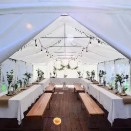 Wnętrze białego namiotu weselnego z drewnianą podłogą, długie stoły z białymi obrusami i jutowymi bieżnikami, drewniane ławy, dekoracje kwiatowe w szklanych wazonach, girlandy z żarówek i trzy...