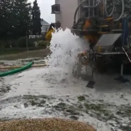 Wiercenie studni: silny strumień wody wydobywa się z otworu w ziemi podczas pracy wiertnicy na tle budynku mieszkalnego i drzew.