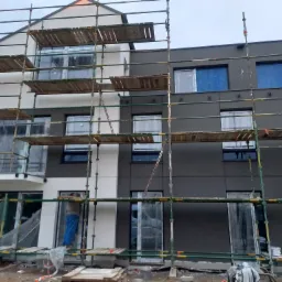 Nowoczesny, dwukolorowy budynek w trakcie prac elewacyjnych, otoczony rusztowaniami, z widocznymi pracownikami przy oknach.