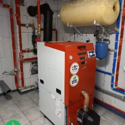 Nowoczesny piec na pellet w kotłowni z biało-szarymi płytkami, widoczne liczne rury instalacji CO w kolorze czerwonym i niebieskim, zbiornik wyrównawczy oraz czarny komin. Piec oznaczony jako 5...