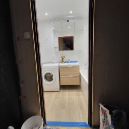 TOMASZ WRZESINSKI PRZEDSIĘBIORSTWO BUDOWLANE TOM-BUD - Widok na wyremontowaną łazienkę z pralką, umywalką z szafką i lustrem, widzianą zza otwartych drzwi, z widocznymi narzędziami i workiem zaprawy na podłodze.