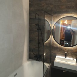 TOMASZ WRZESINSKI PRZEDSIĘBIORSTWO BUDOWLANE TOM-BUD - Nowoczesna łazienka z beżowymi i brązowymi płytkami, czarną armaturą i okrągłym lustrem z podświetleniem. Widoczne odbicie osoby robiącej zdjęcie.