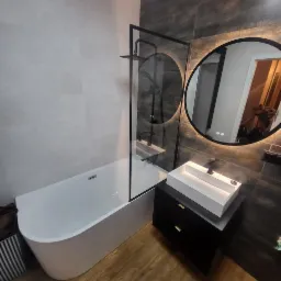 Nowoczesna łazienka z wolnostojącą wanną, szklanym parawanem i okrągłym lustrem z podświetleniem LED. Ciemne płytki kontrastują z bielą armatury.