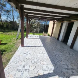 Zadaszony taras z nową, wzorzystą terakotą na podłodze, drewniane słupy i biały sufit, drabina oparta o słup na zewnątrz.