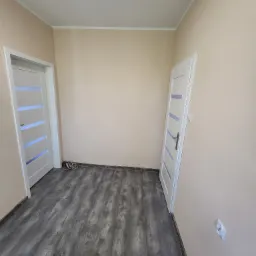 Wnętrze pokoju z nową szarą podłogą imitującą drewno, dwiema białymi drzwiami z przeszkleniami i beżowymi ścianami; widoczny wystający kabel elektryczny przy jednej ze ścian.