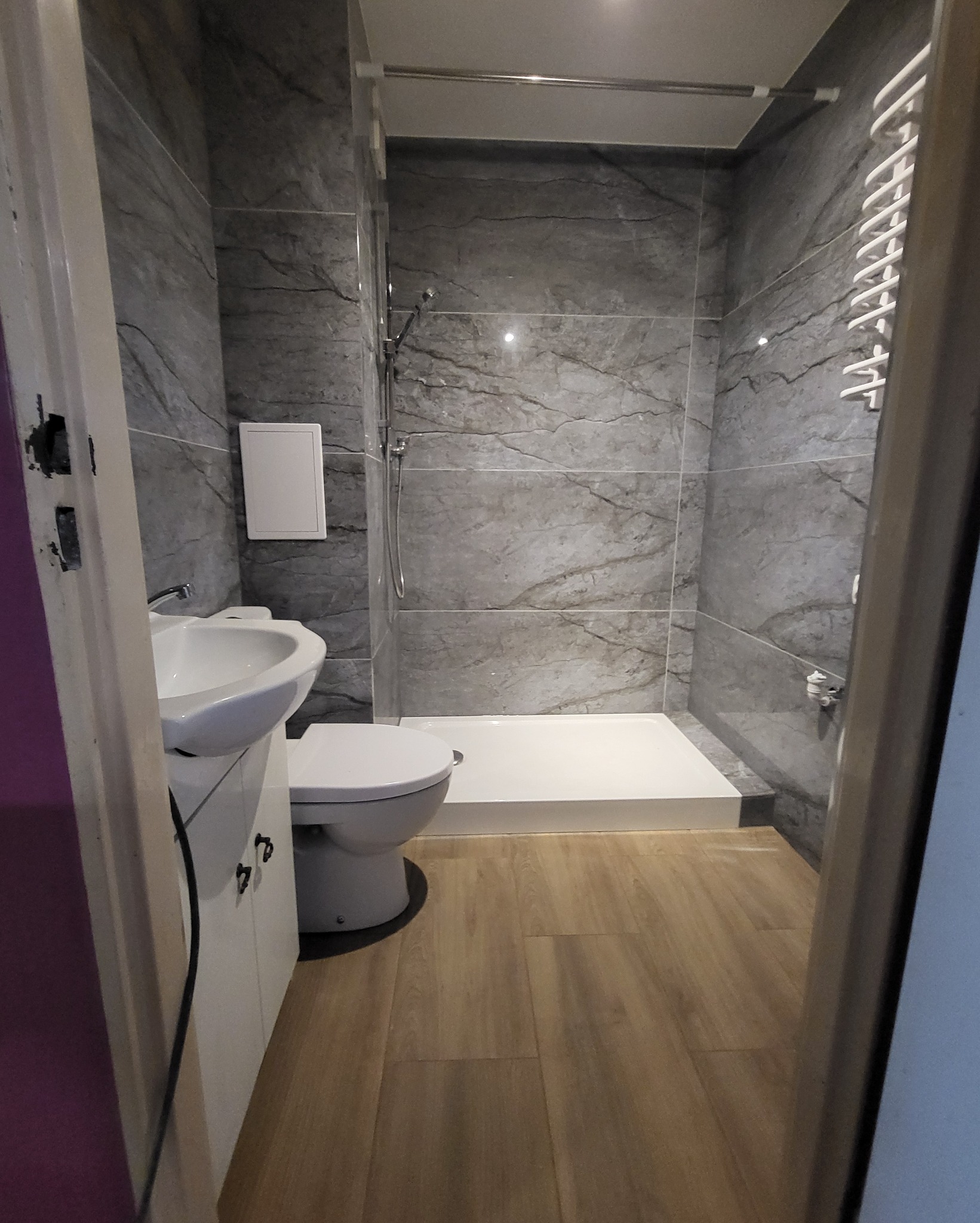 Wnętrze wyremontowanej łazienki z kabiną prysznicową typu walk-in, umywalką z szafką, toaletą i grzejnikiem drabinkowym, wykończonej szarymi płytkami imitującymi marmur i panelami podłogowymi...