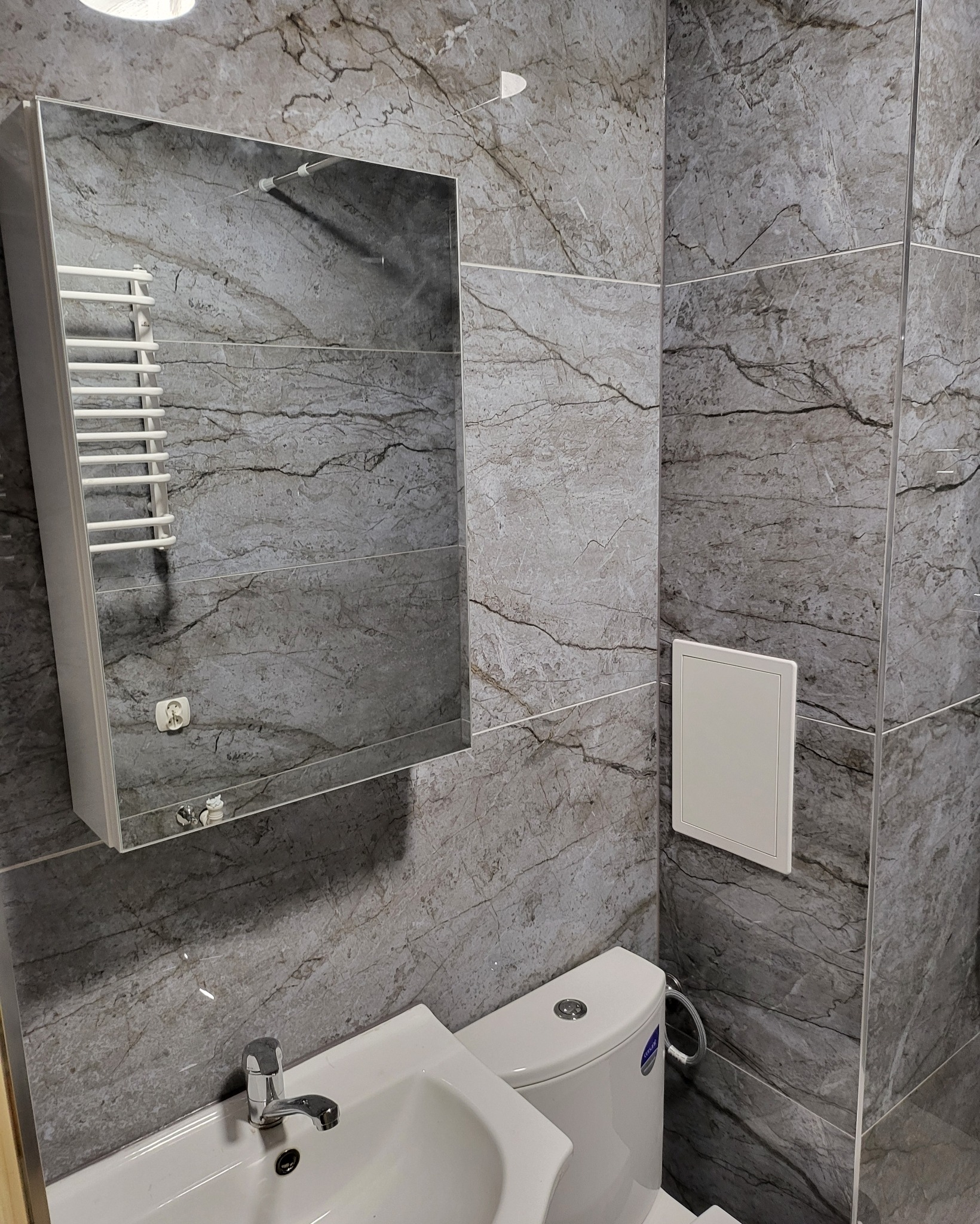 Szara łazienka z płytkami imitującymi marmur, umywalką, toaletą i grzejnikiem drabinkowym odbijającym się w lustrze.
