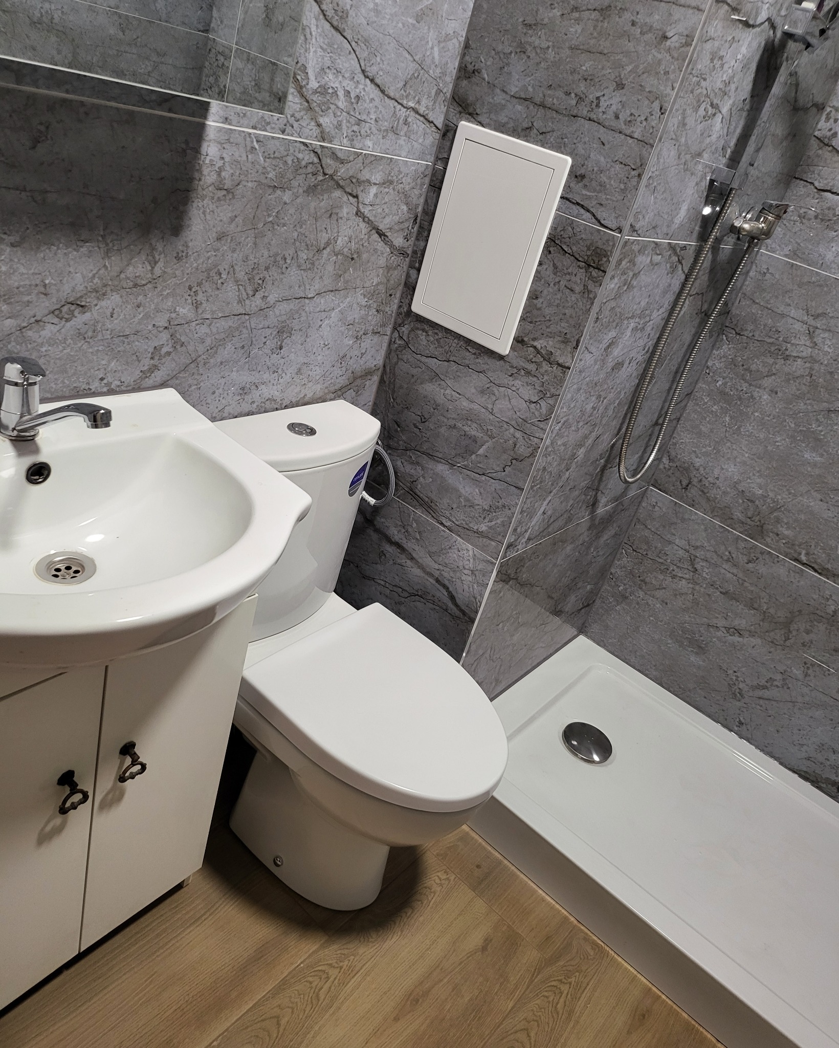 Nowoczesna łazienka z szarymi płytkami imitującymi marmur, białą umywalką z szafką z ozdobnymi uchwytami, toaletą i brodzikiem z odpływem liniowym.