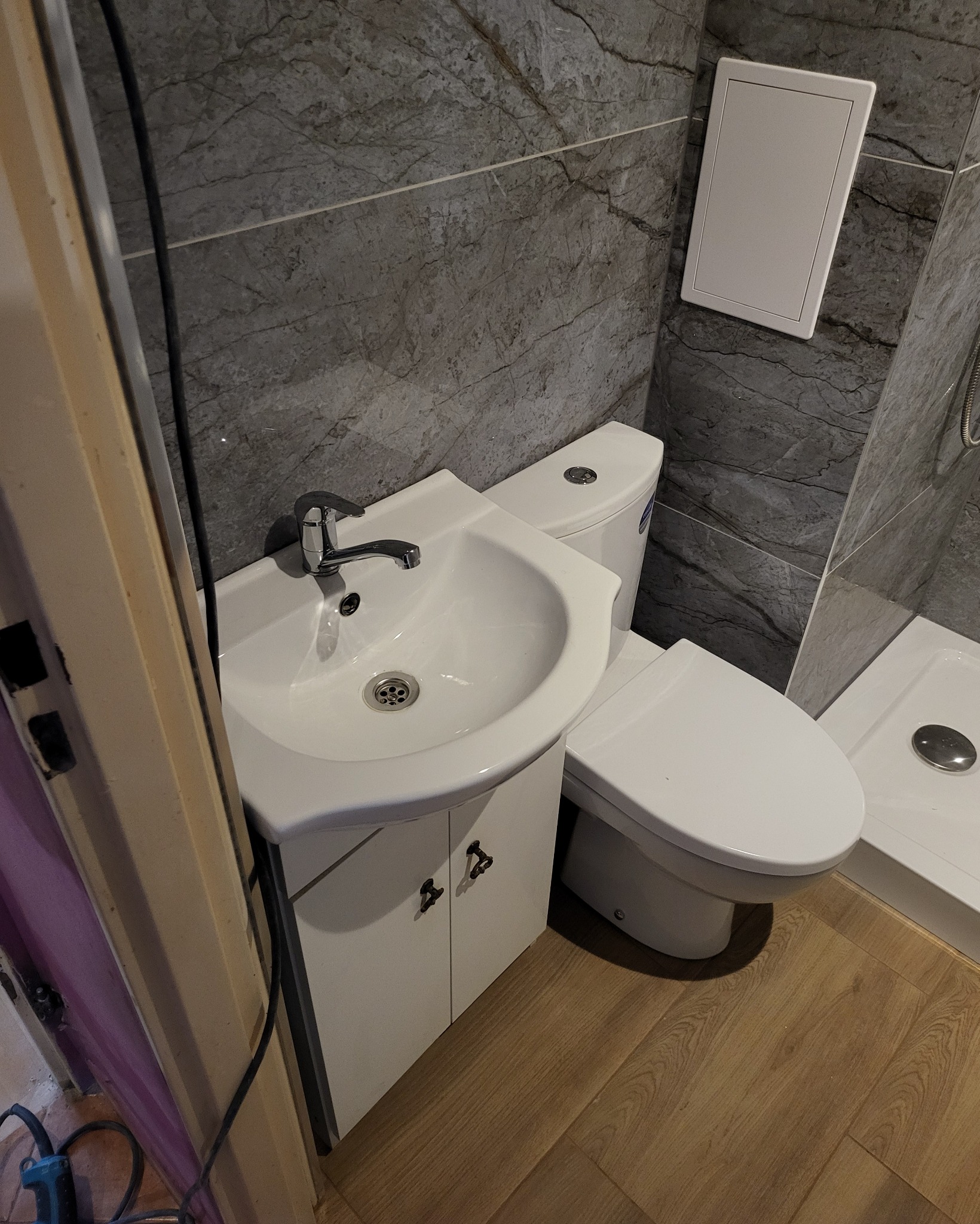 Ukończona biała umywalka z szafką, toaleta i brodzik z szarą marmurową okładziną ścienną oraz ukrytymi drzwiczkami rewizyjnymi, widoczne drewnopodobne panele podłogowe.