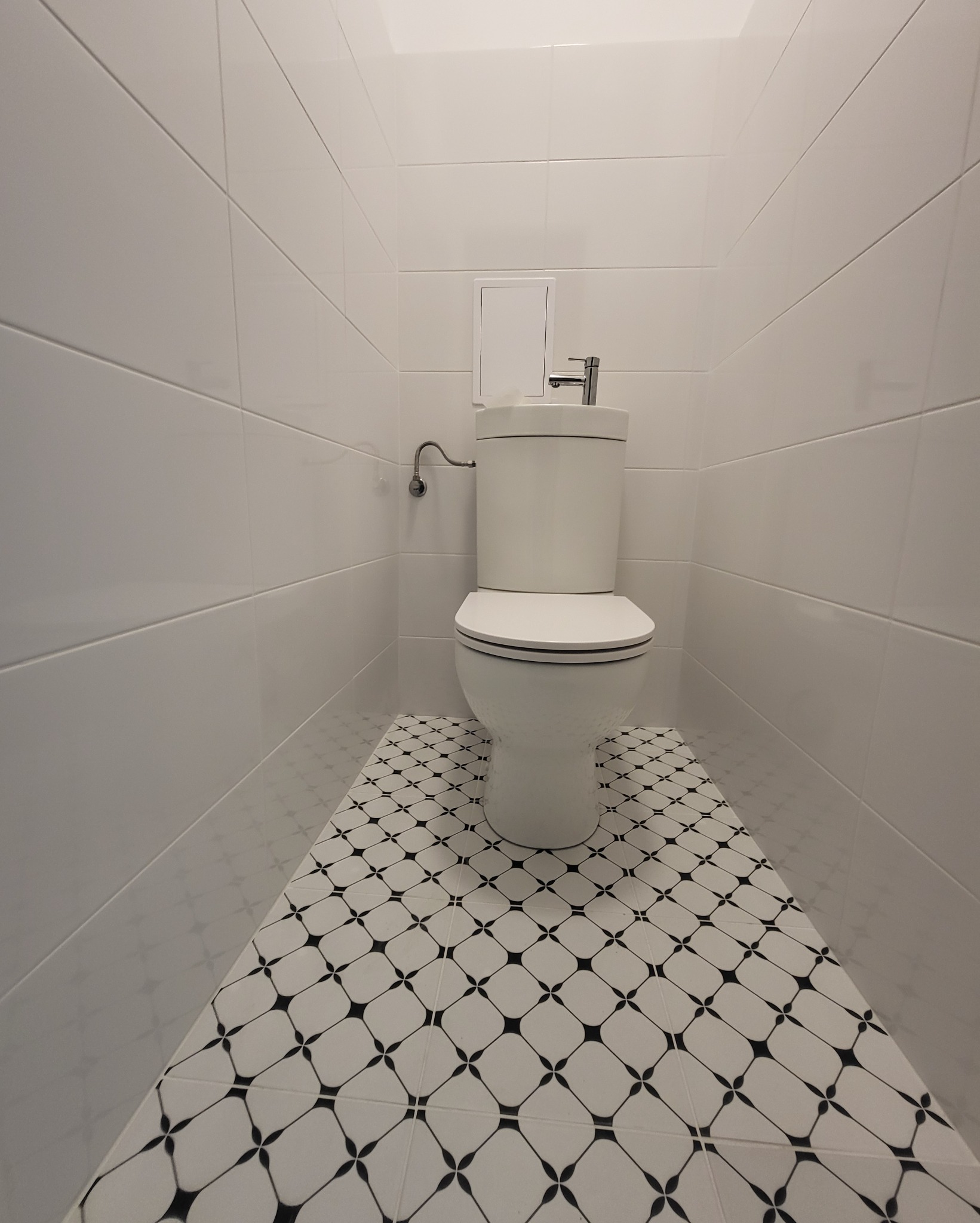 Kompaktowa toaleta z białą ceramiką, chromowaną armaturą i ozdobnymi płytkami podłogowymi w wąskiej, wykafelkowanej przestrzeni.