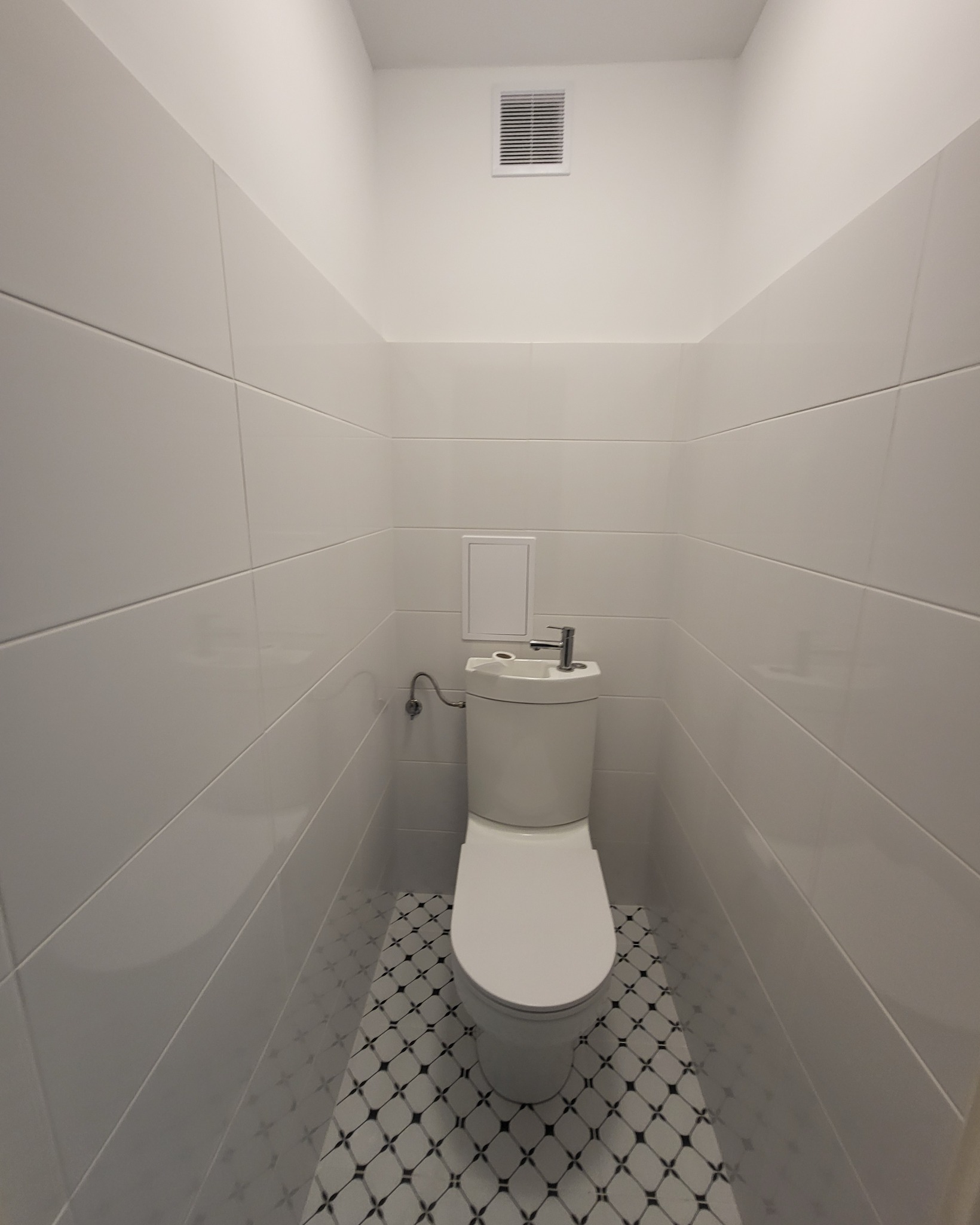 Kompaktowa toaleta z umywalką na spłuczce, białe, błyszczące płytki na ścianach ułożone poziomo, podłoga w biało-czarną mozaikę, wentylacja na suficie.