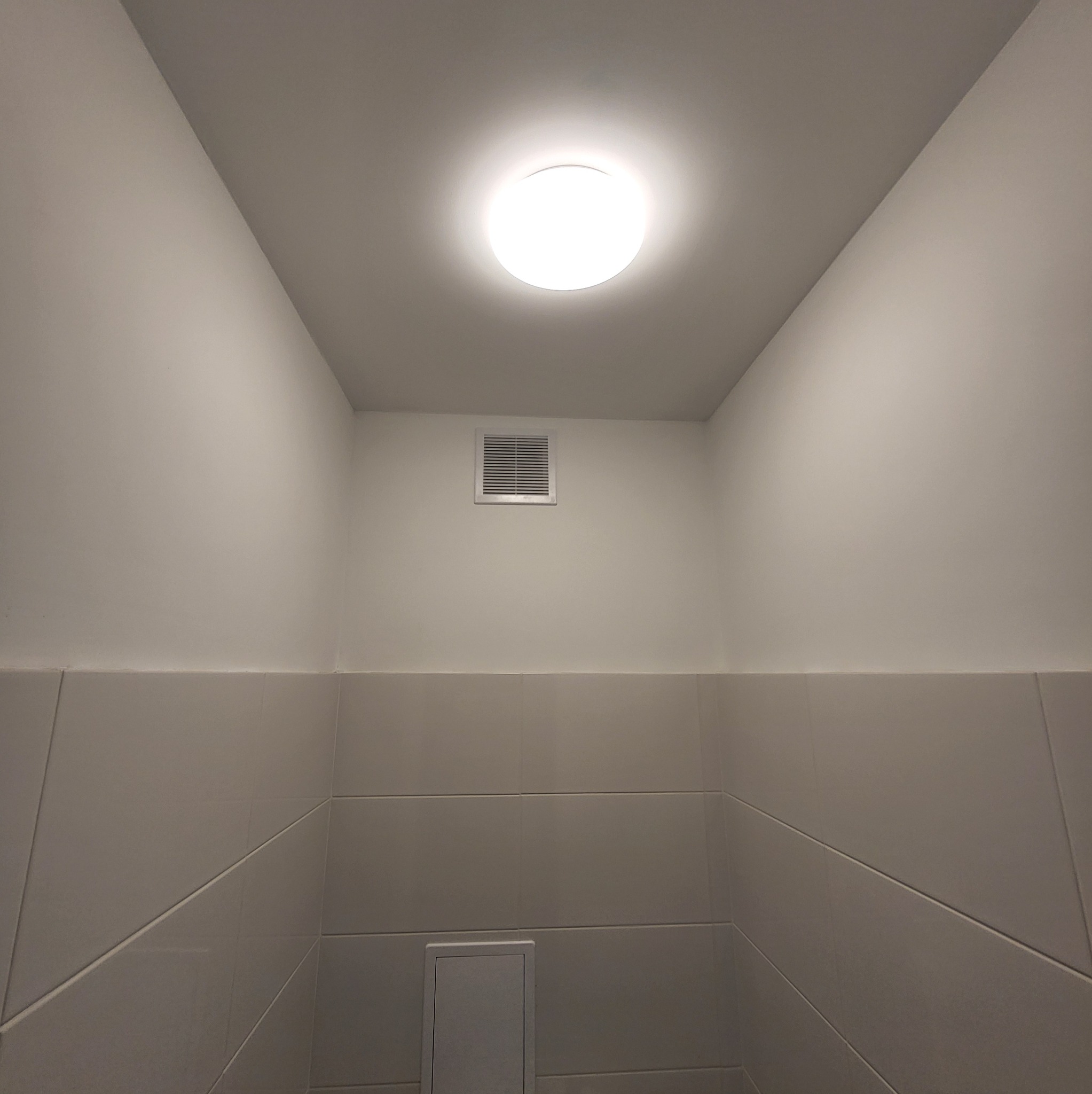 Wnętrze wykończonej łazienki z szarymi płytkami, widoczna kratka wentylacyjna i okrągła lampa sufitowa.