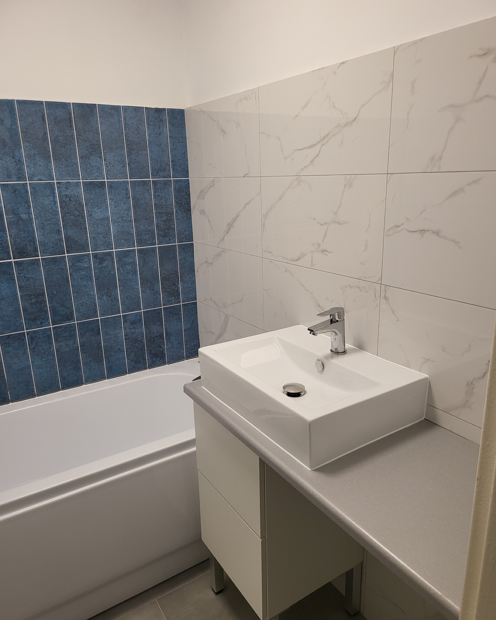 Nowoczesna łazienka po remoncie z prostokątną umywalką nablatową, baterią chromowaną, białą wanną i ścianami wyłożonymi niebieskimi płytkami wertykalnymi oraz białymi płytkami z szarym marmurkowym...