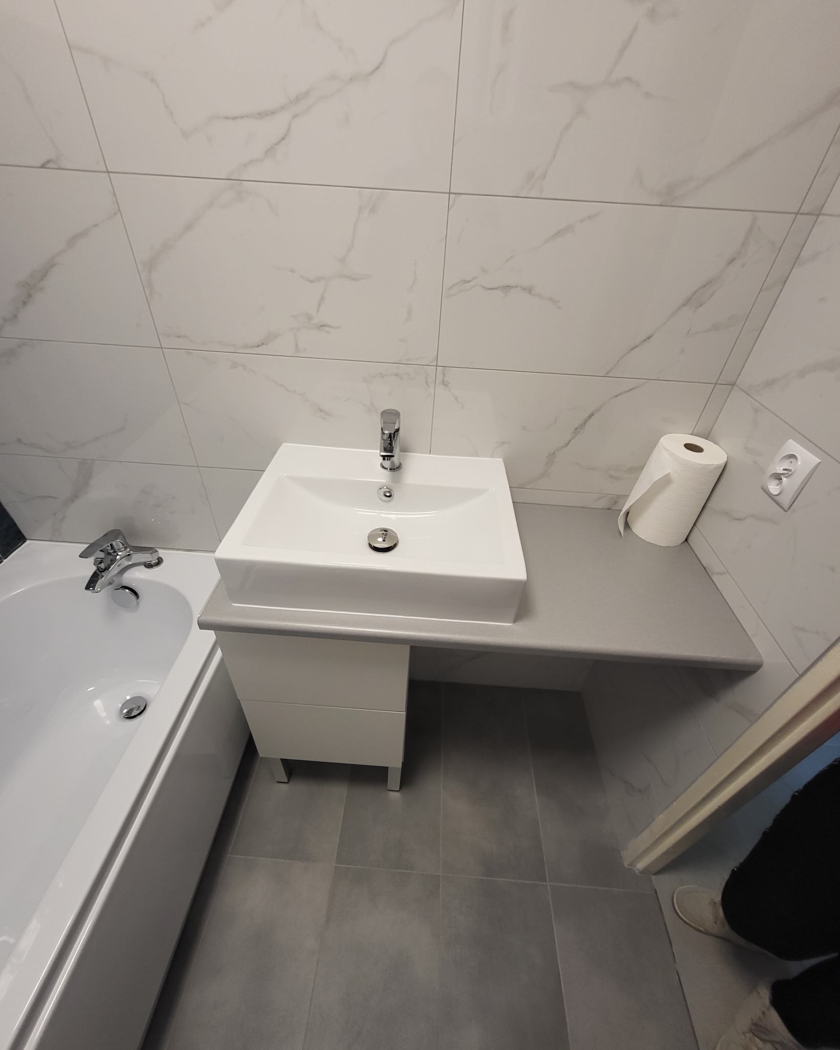 Nowoczesna łazienka z białą umywalką nablatową na szarym blacie, białe płytki imitujące marmur na ścianach, szara podłoga, obok wanna i rolka papieru toaletowego.