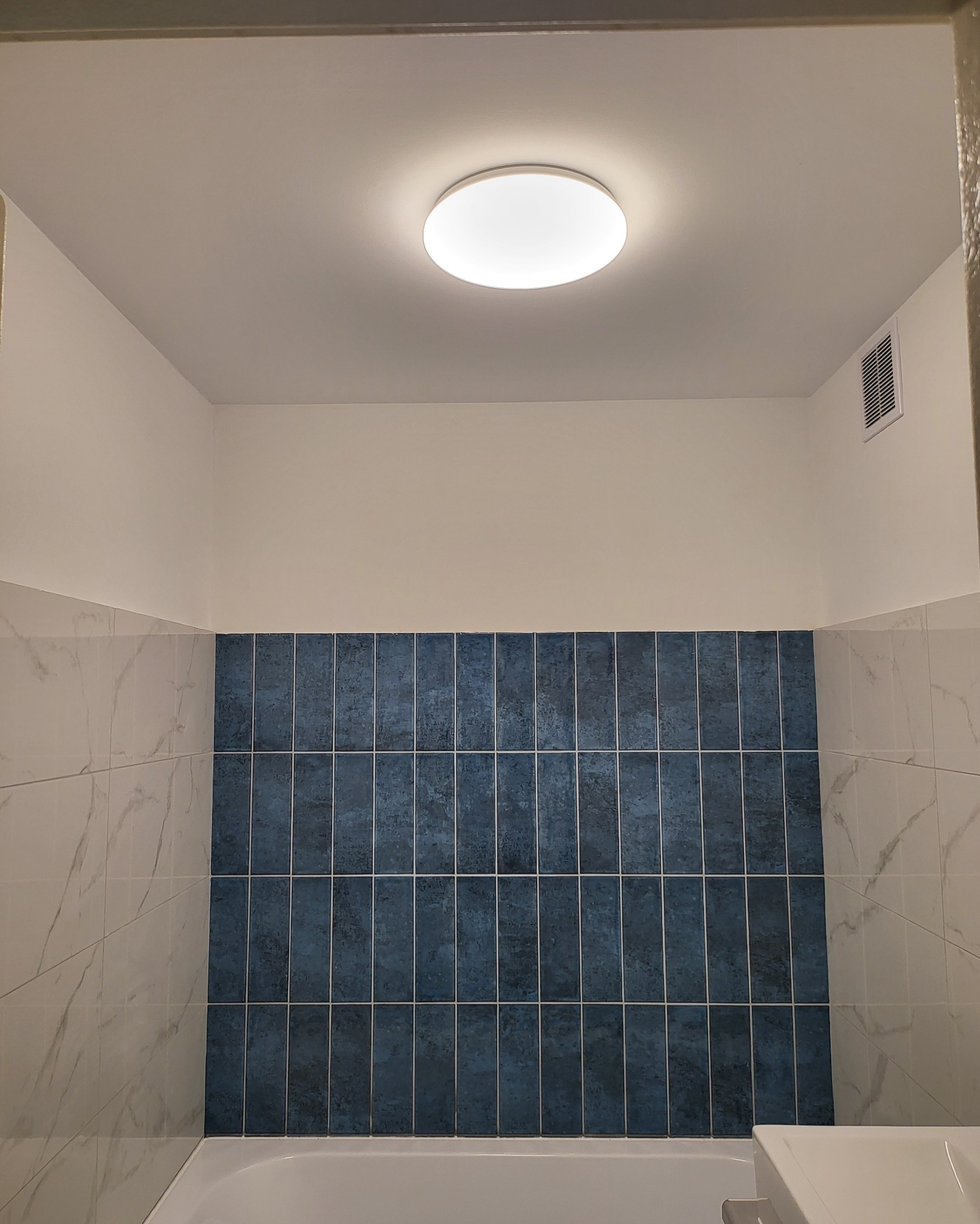 Wykończona łazienka z niebieskimi płytkami ułożonymi pionowo, marmurowe płytki po bokach, okrągła lampa sufitowa i kratka wentylacyjna.