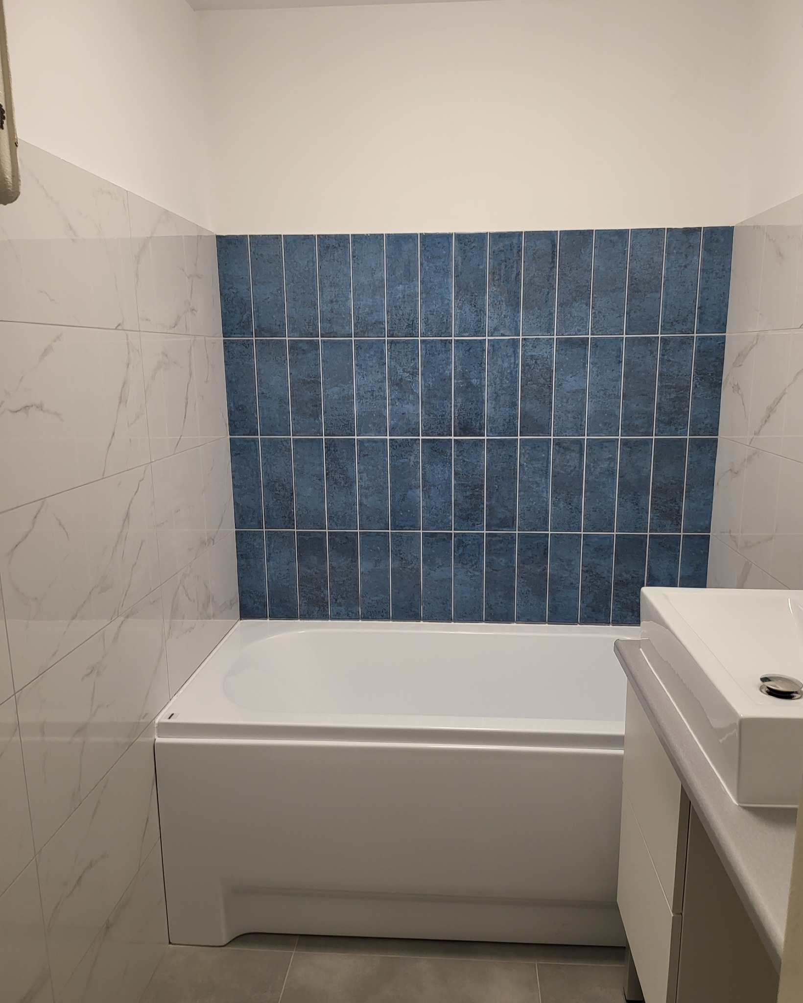 Nowoczesna łazienka po remoncie: biała wanna zabudowana z niebieskimi płytkami ułożonymi pionowo na ścianie, obok umywalka z szafką, marmurowe płytki na ścianach.