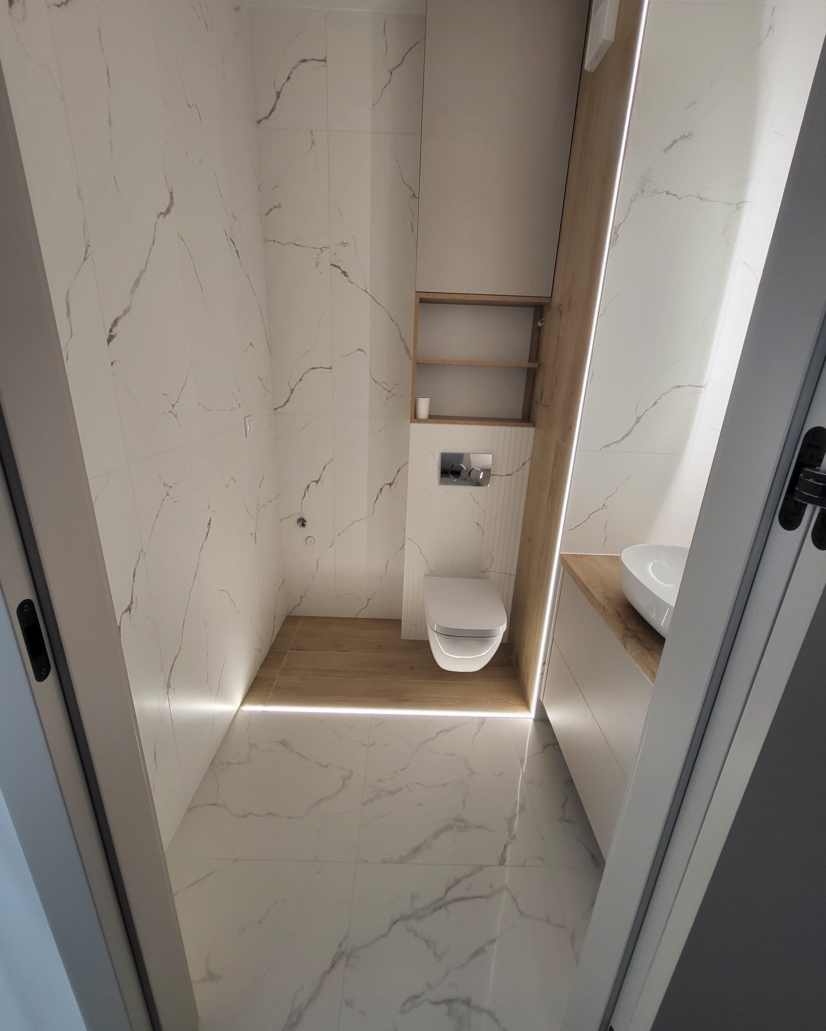 Wykończona łazienka w bieli i drewnie: marmurowe płytki, podwieszana toaleta, szafki z półkami i oświetlenie LED podłogowe, widok zza uchylonych drzwi.