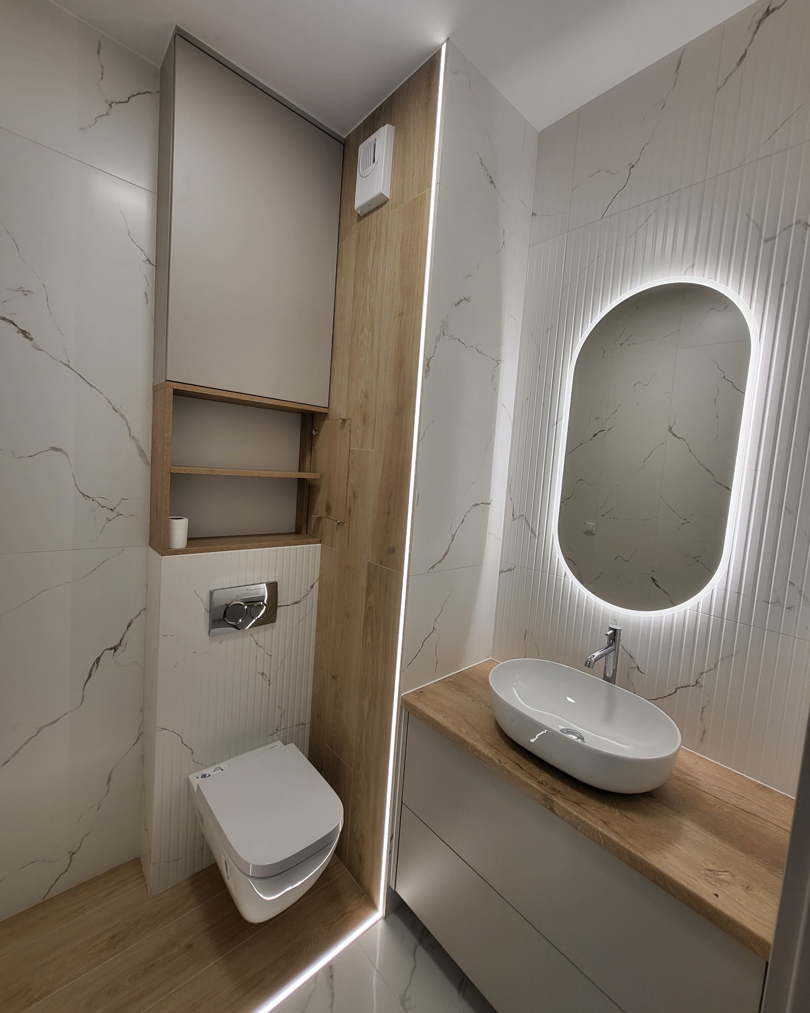 Nowoczesna łazienka z podświetlanym lustrem, umywalką nablatową i wiszącą toaletą. Płytki imitujące marmur i drewno, oświetlenie LED.