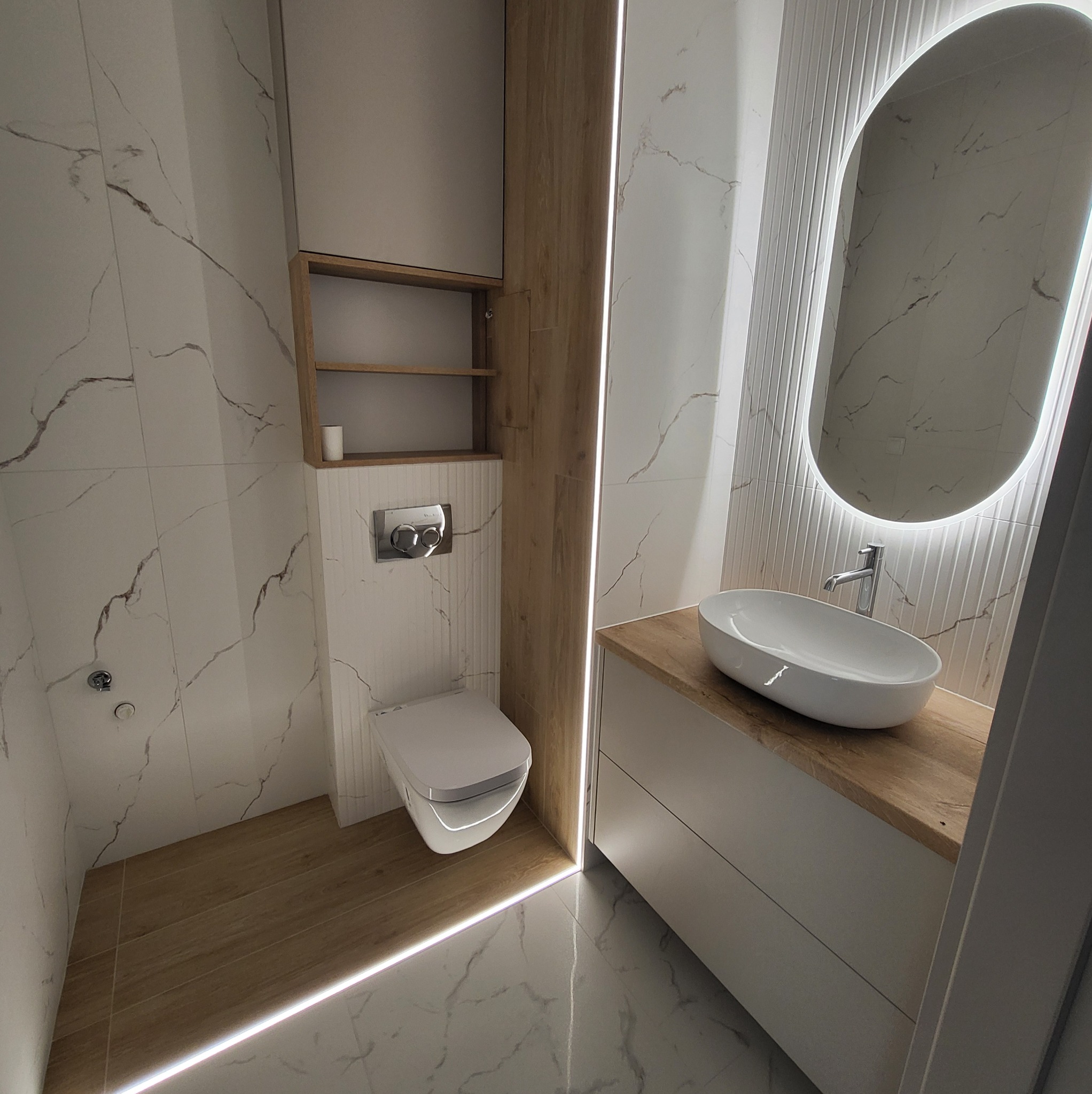 Nowoczesna łazienka po remoncie: białe płytki z szarymi żyłkami, podświetlane lustro owalne, drewniane akcenty i podwieszana toaleta. Estetyczne wykończenie.
