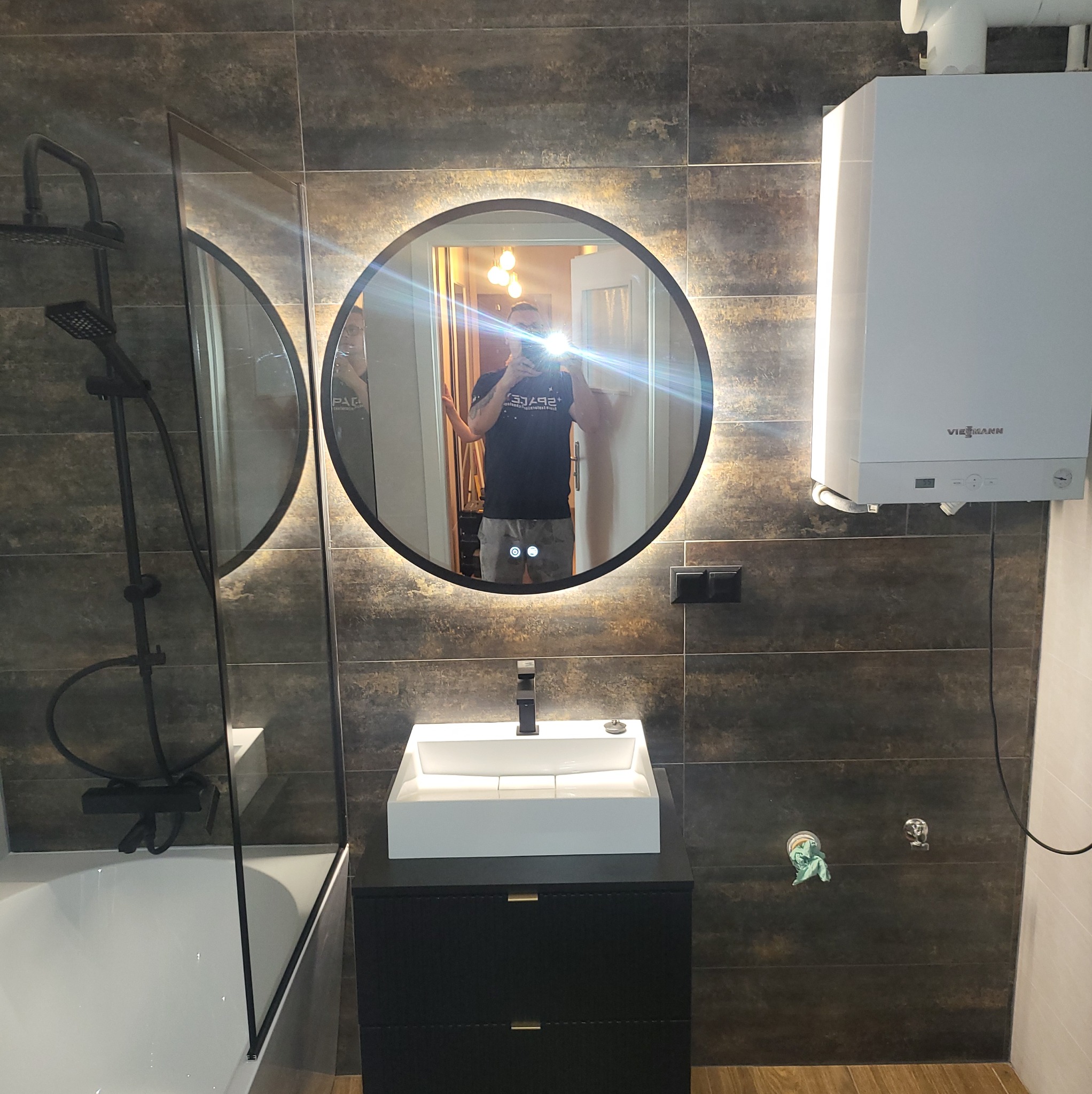 Nowoczesna łazienka z okrągłym lustrem LED, białą umywalką na czarnej szafce i kabiną prysznicową. Ciemne płytki na ścianach i jasna podłoga.