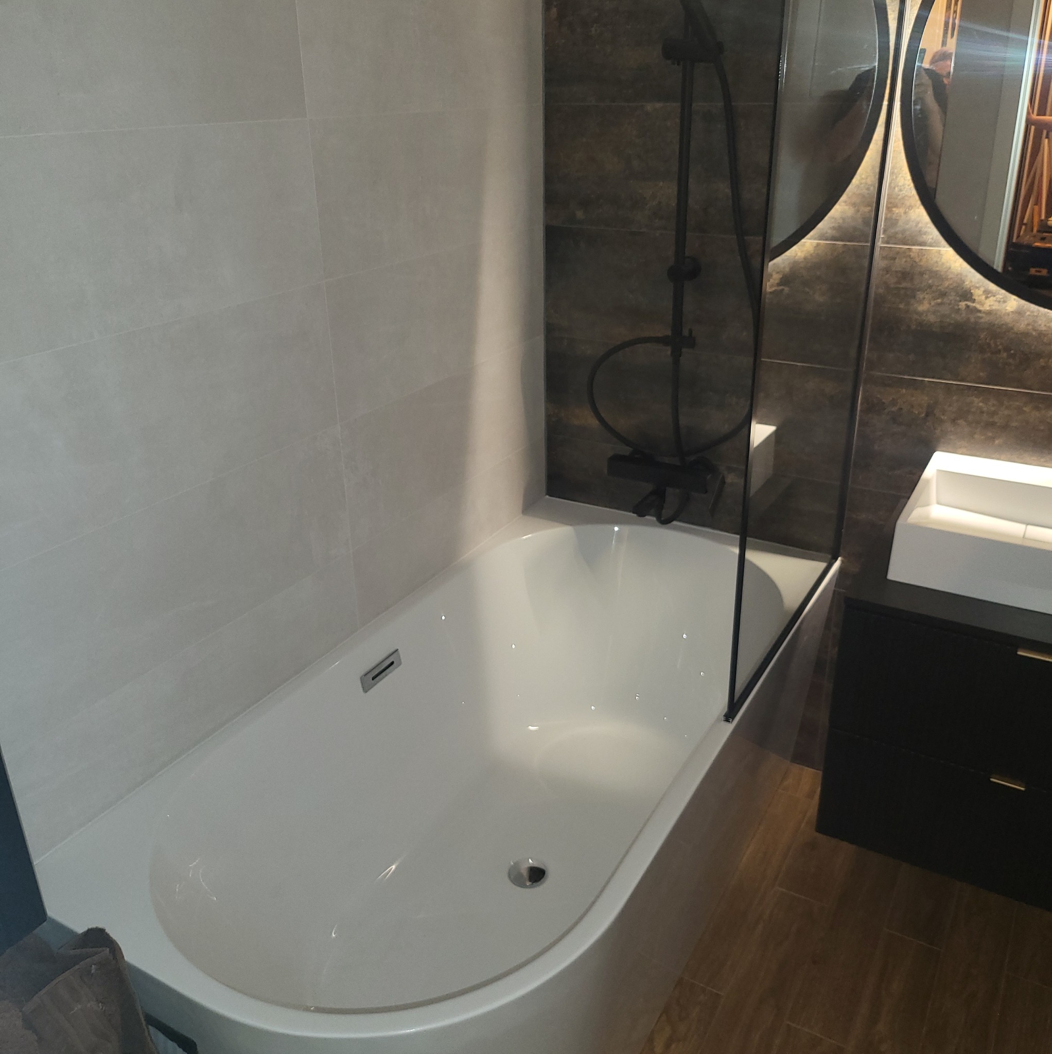 Nowoczesna łazienka z wanną, czarną armaturą i dużym lustrem z podświetleniem LED. Jasne płytki na ścianach kontrastują z ciemnymi elementami wyposażenia.