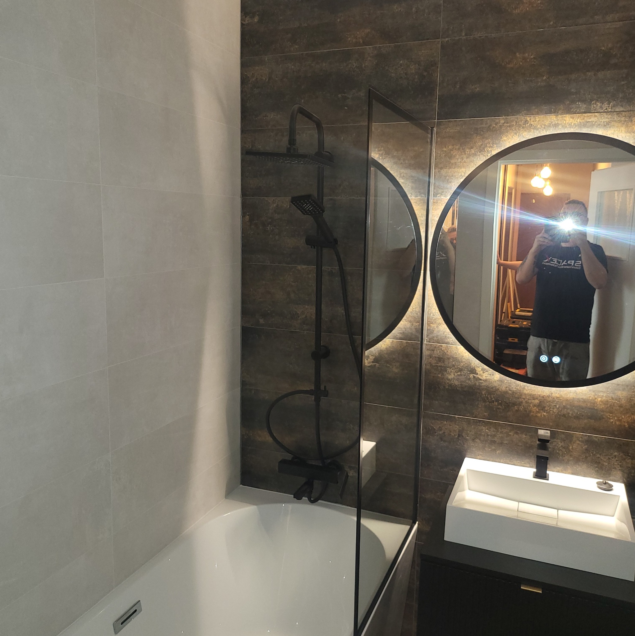 Nowoczesna łazienka z beżowymi i brązowymi płytkami, czarną armaturą i okrągłym lustrem z podświetleniem. Widoczne odbicie osoby robiącej zdjęcie.