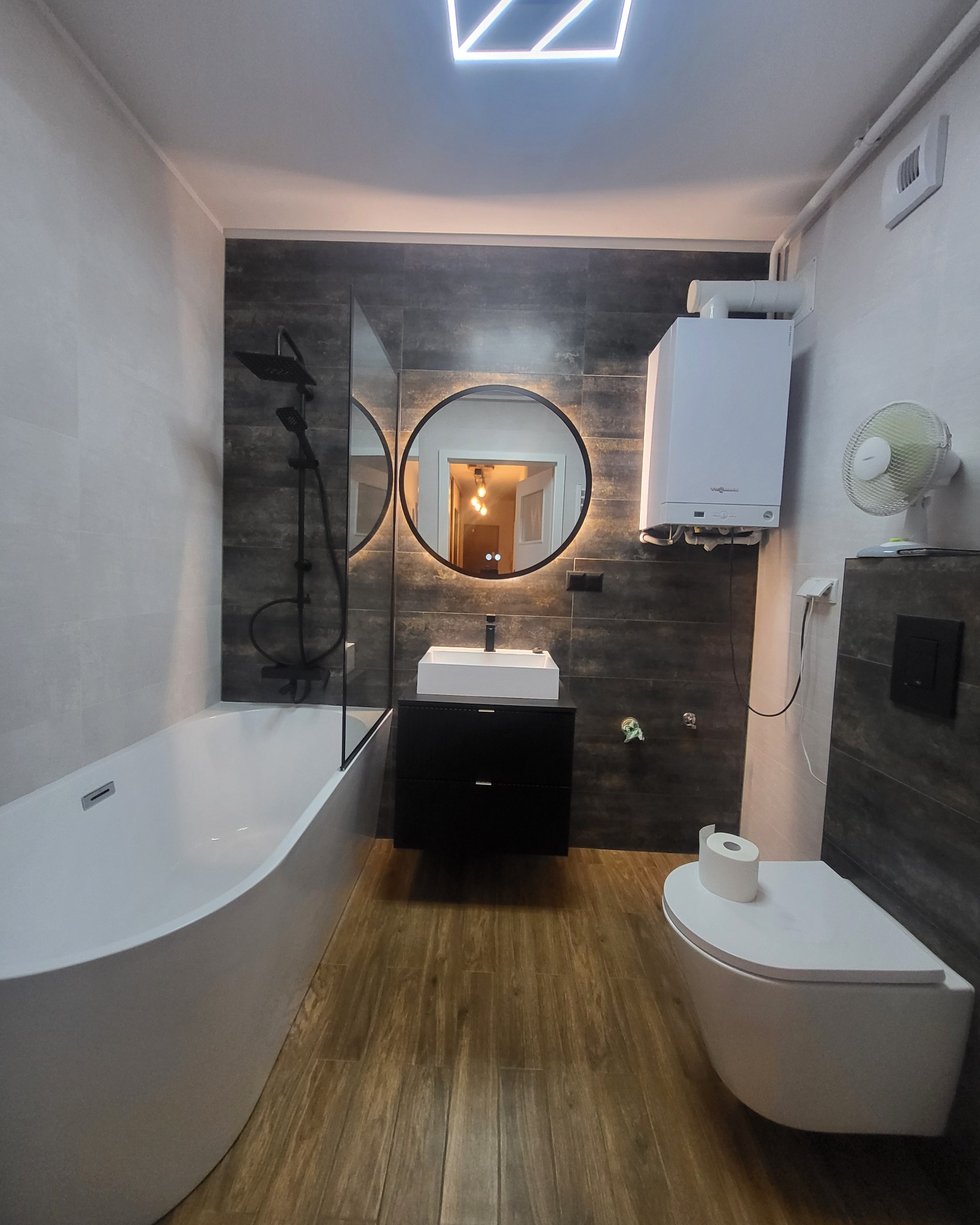 Nowoczesna łazienka z wanną, kabiną prysznicową typu walk-in, okrągłym lustrem z podświetleniem i drewnopodobną podłogą. Widoczny bojler i wentylator.