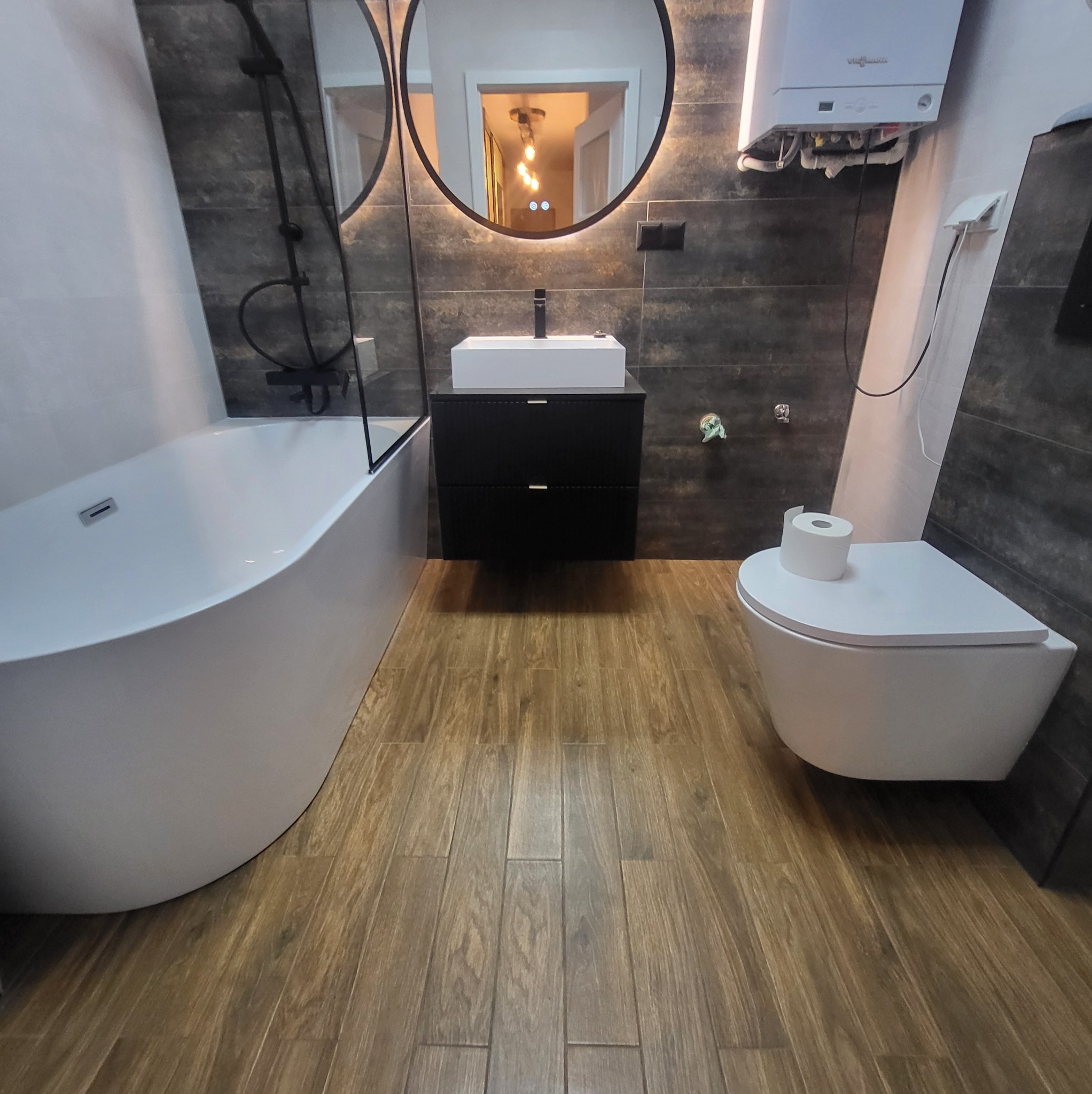 Nowoczesna łazienka z drewnopodobną podłogą, białą wanną i umywalką na czarnej szafce. Na ścianach ciemne płytki imitujące kamień i okrągłe lustro z podświetleniem LED.