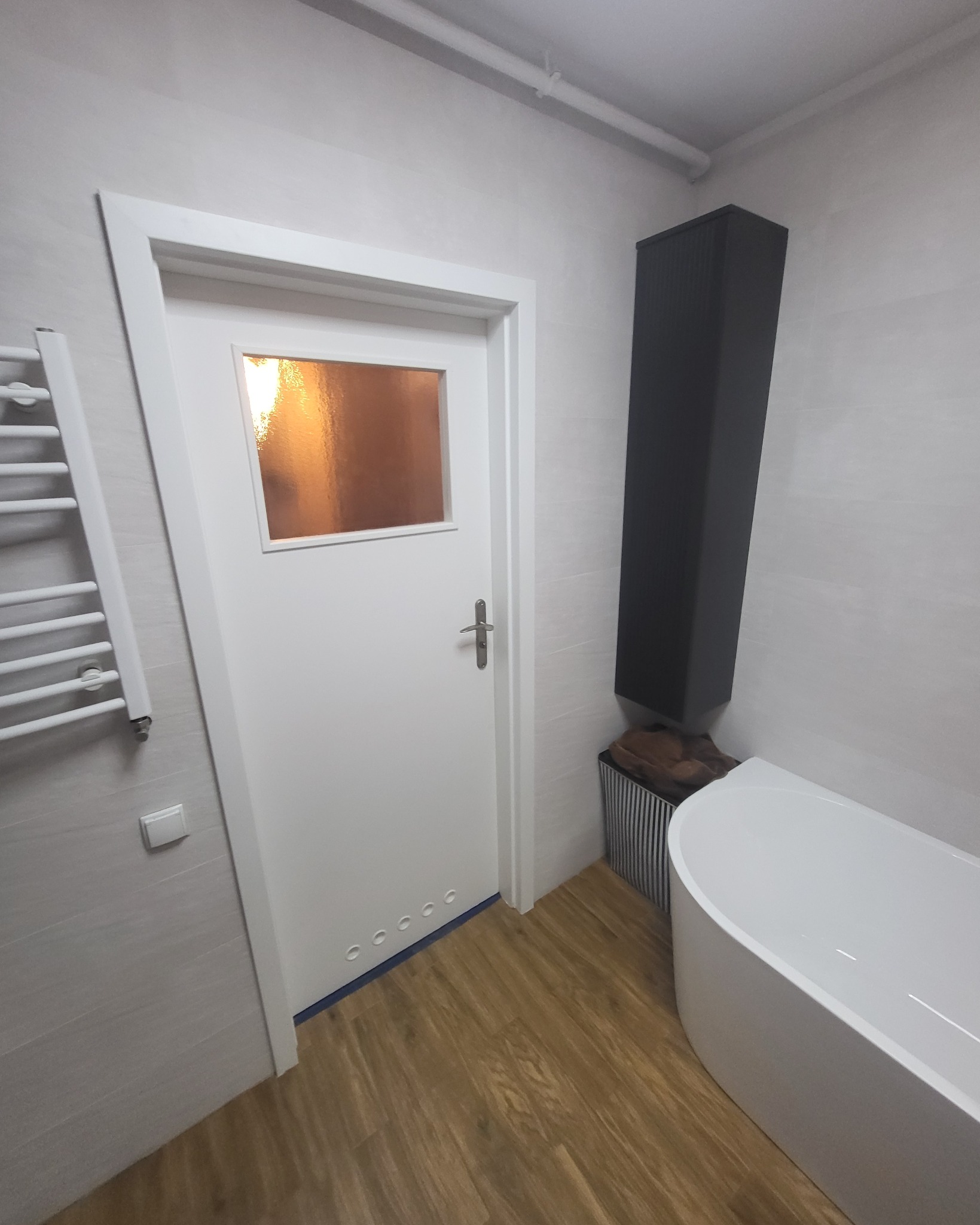 Nowoczesna łazienka po remoncie: biała wanna wolnostojąca, drzwi z szybą, grzejnik drabinkowy i drewnopodobne panele podłogowe. Minimalistyczny design.