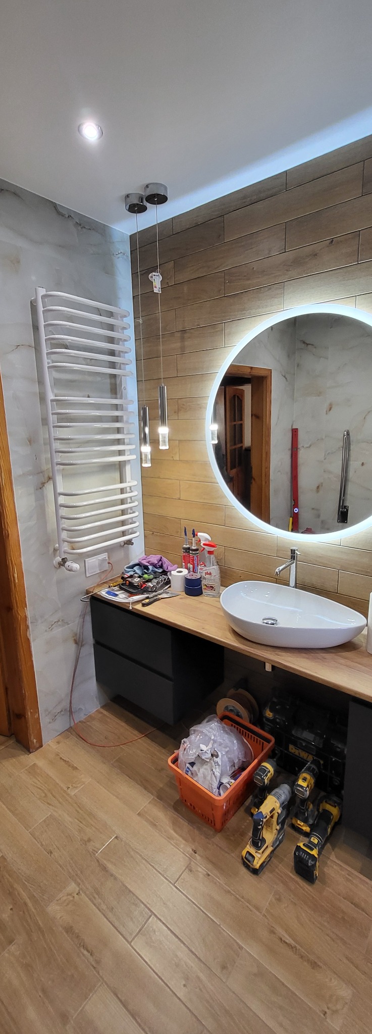 Nowoczesna łazienka w trakcie wykańczania: okrągłe lustro z podświetleniem, drewnopodobne płytki, biały grzejnik drabinkowy i narzędzia glazurnicze.