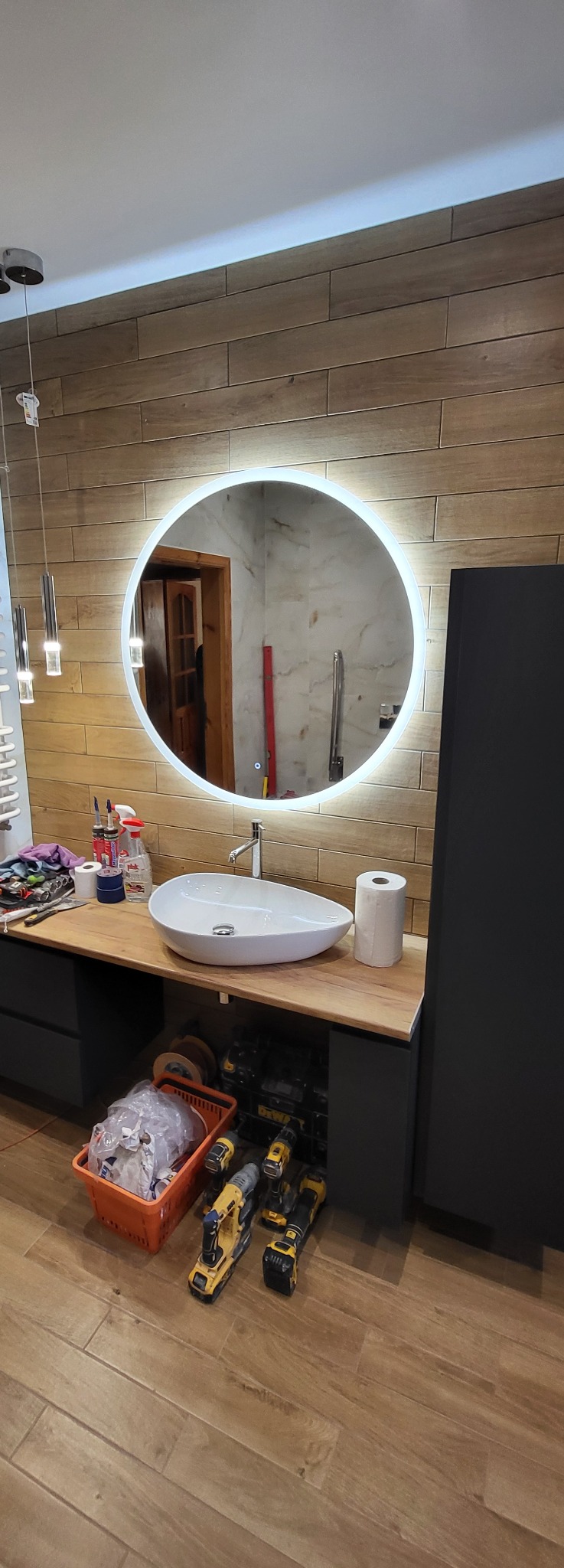 Nowoczesna łazienka po remoncie: drewnopodobne płytki, okrągłe lustro z podświetleniem LED, designerska umywalka nablatowa i czarne szafki. Widoczne narzędzia.