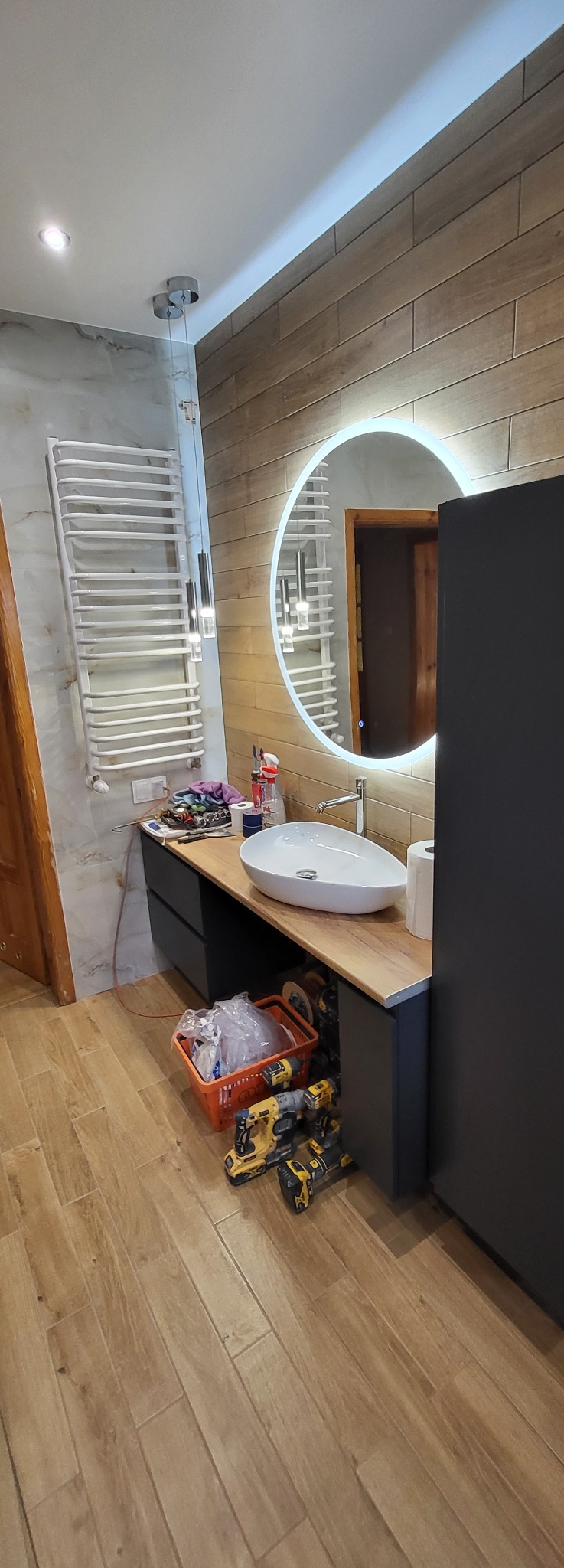 Nowoczesna łazienka po remoncie: drewnopodobne płytki, okrągłe lustro z podświetleniem LED, biała umywalka nablatowa i grzejnik drabinkowy. Widoczne narzędzia.