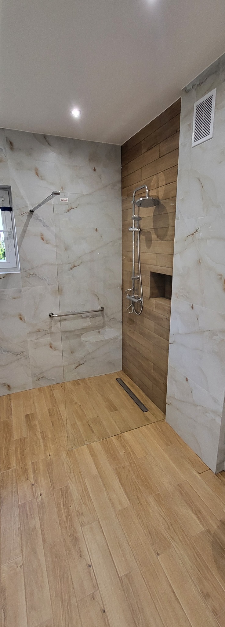 Nowoczesna łazienka po remoncie: marmurowe płytki ścienne, drewnopodobna podłoga, szklana kabina prysznicowa z deszczownicą i odpływem liniowym.
